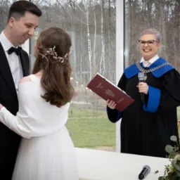 Uśmiechnięta urzędniczka stanu cywilnego w todze z mikrofonem, para młoda w eleganckich strojach podczas ceremonii ślubnej na tle okna z widokiem na drzewa.