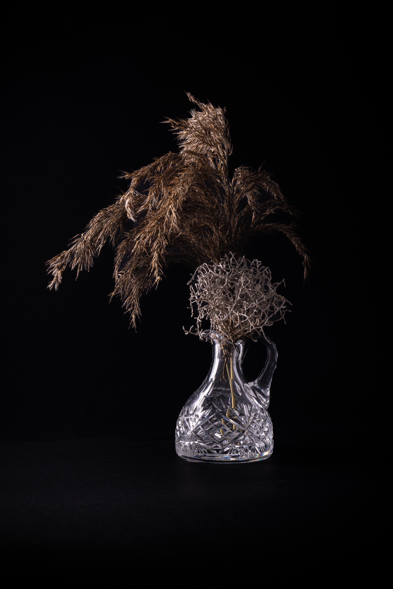 Dekoracyjny, kryształowy wazon z suszonymi trawami na czarnym tle, wykonany w Gdańsku.