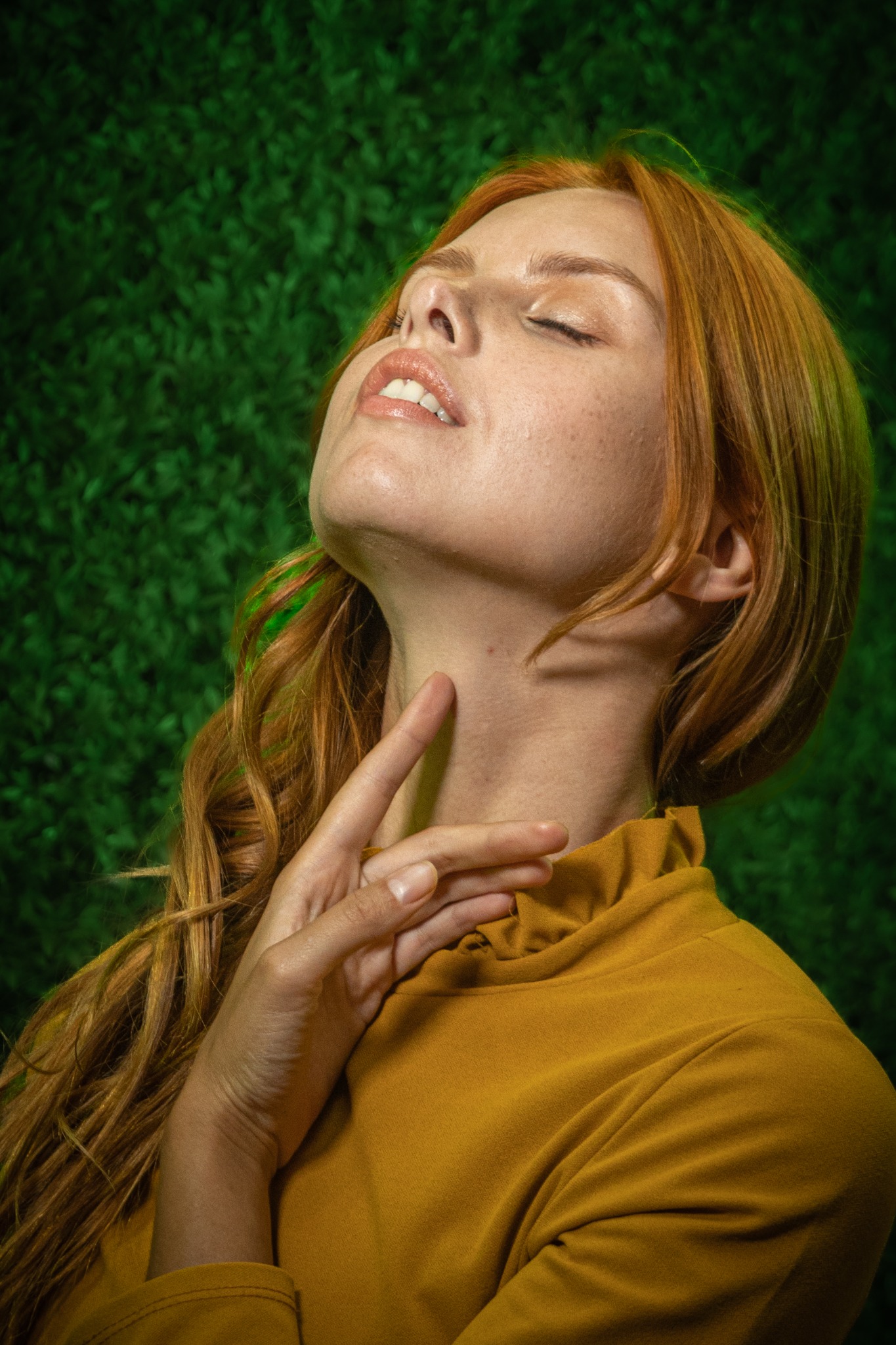 Portret kobiety z rudymi włosami, zamkniętymi oczami i ręką na szyi, na tle zielonej trawy