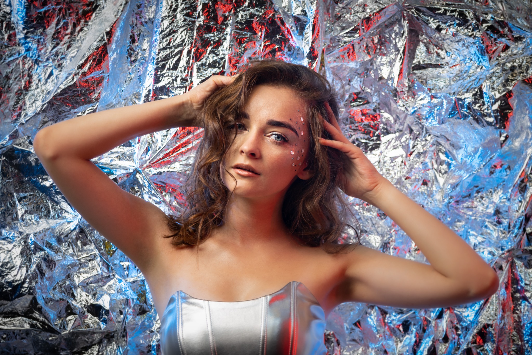 Portret kobiety z błyszczącym makijażem i srebrnym topem na tle folii aluminiowej z czerwono-niebieskim oświetleniem.