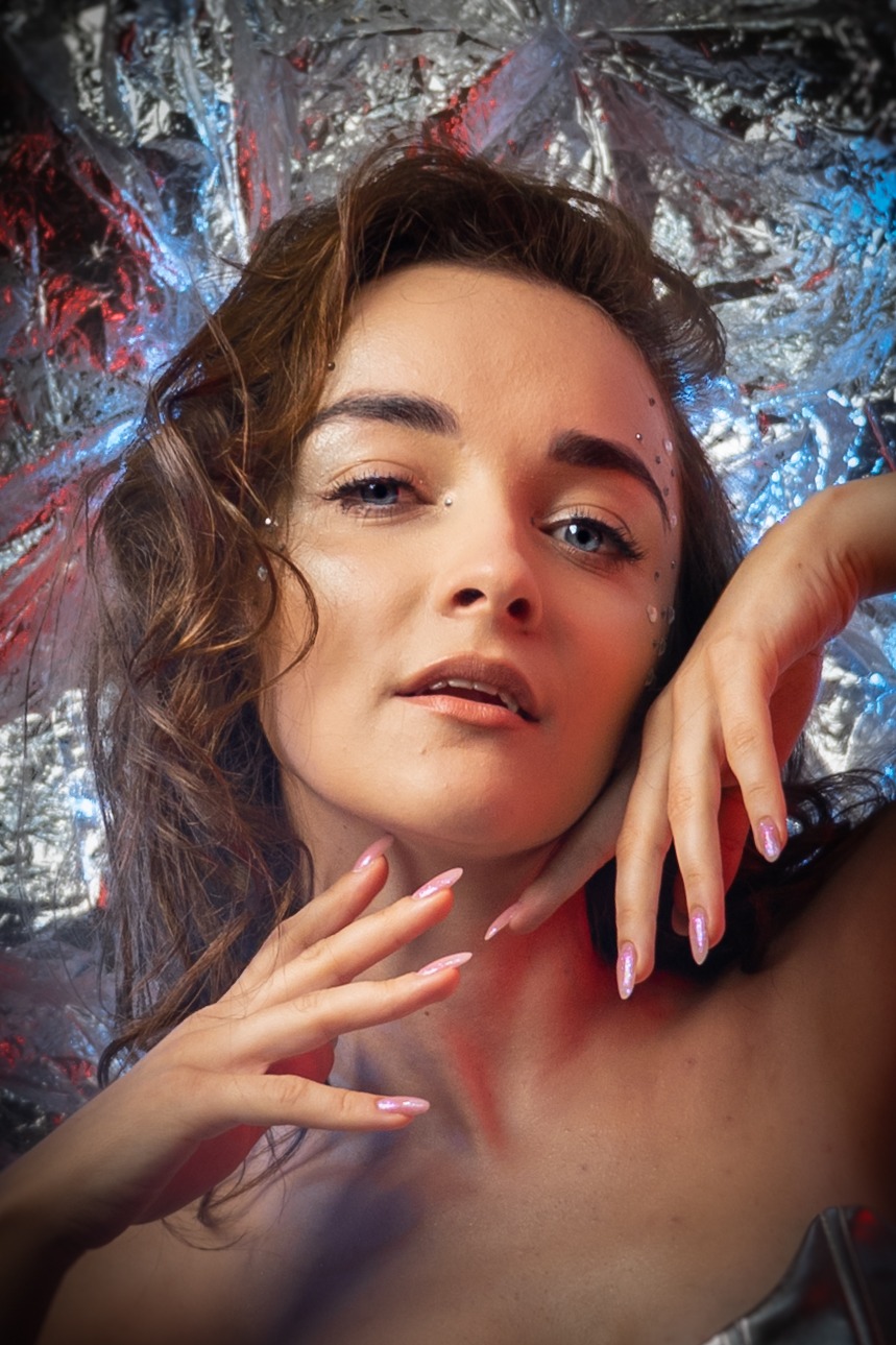 Portret kobiety z błyszczącymi kryształkami na twarzy i opalizującym manicure na tle srebrnej folii.