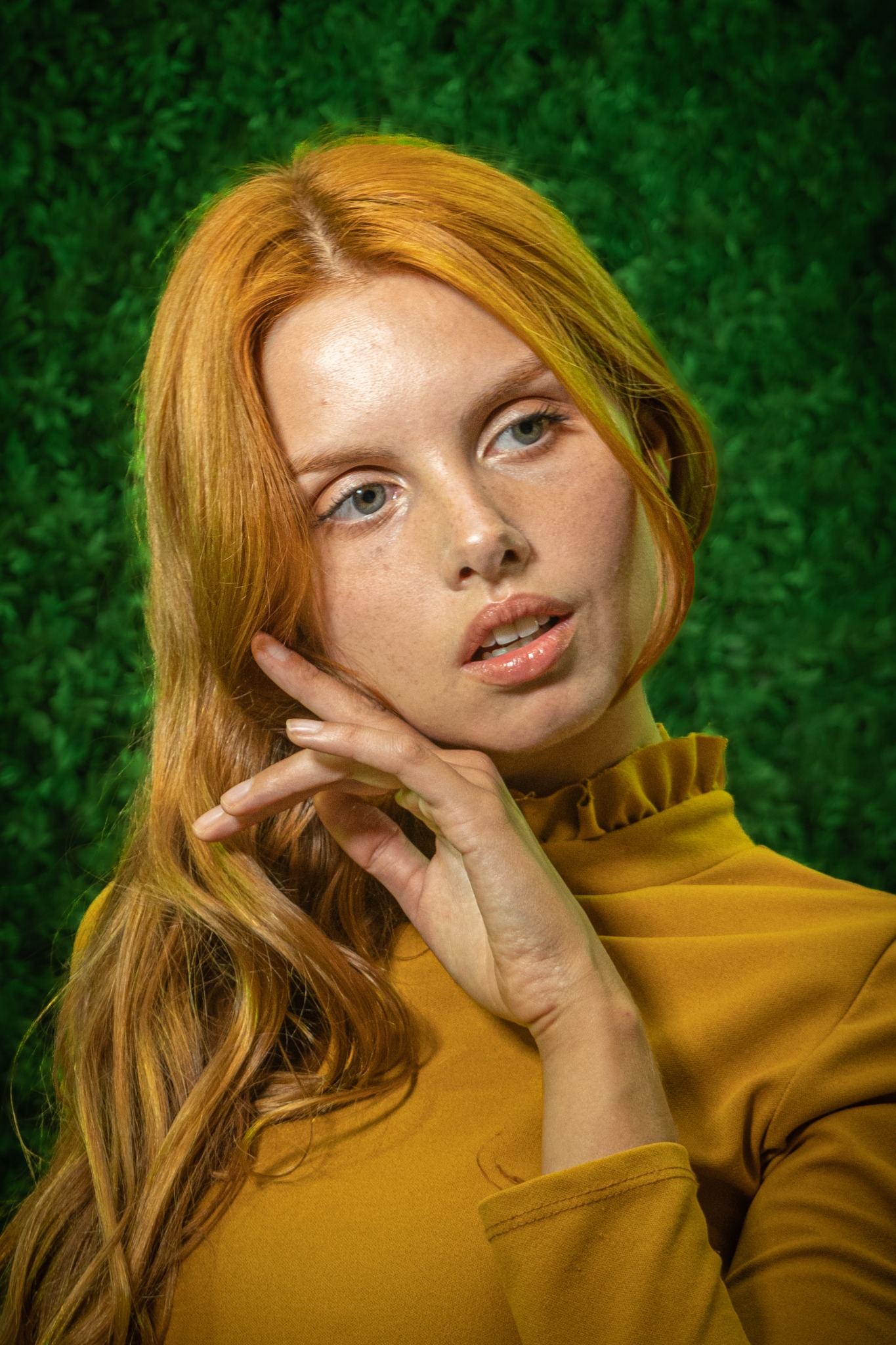 Portret kobiety z rudymi włosami, w musztardowej bluzce z falbanką, pozującej na tle ściany z zielonych roślin.