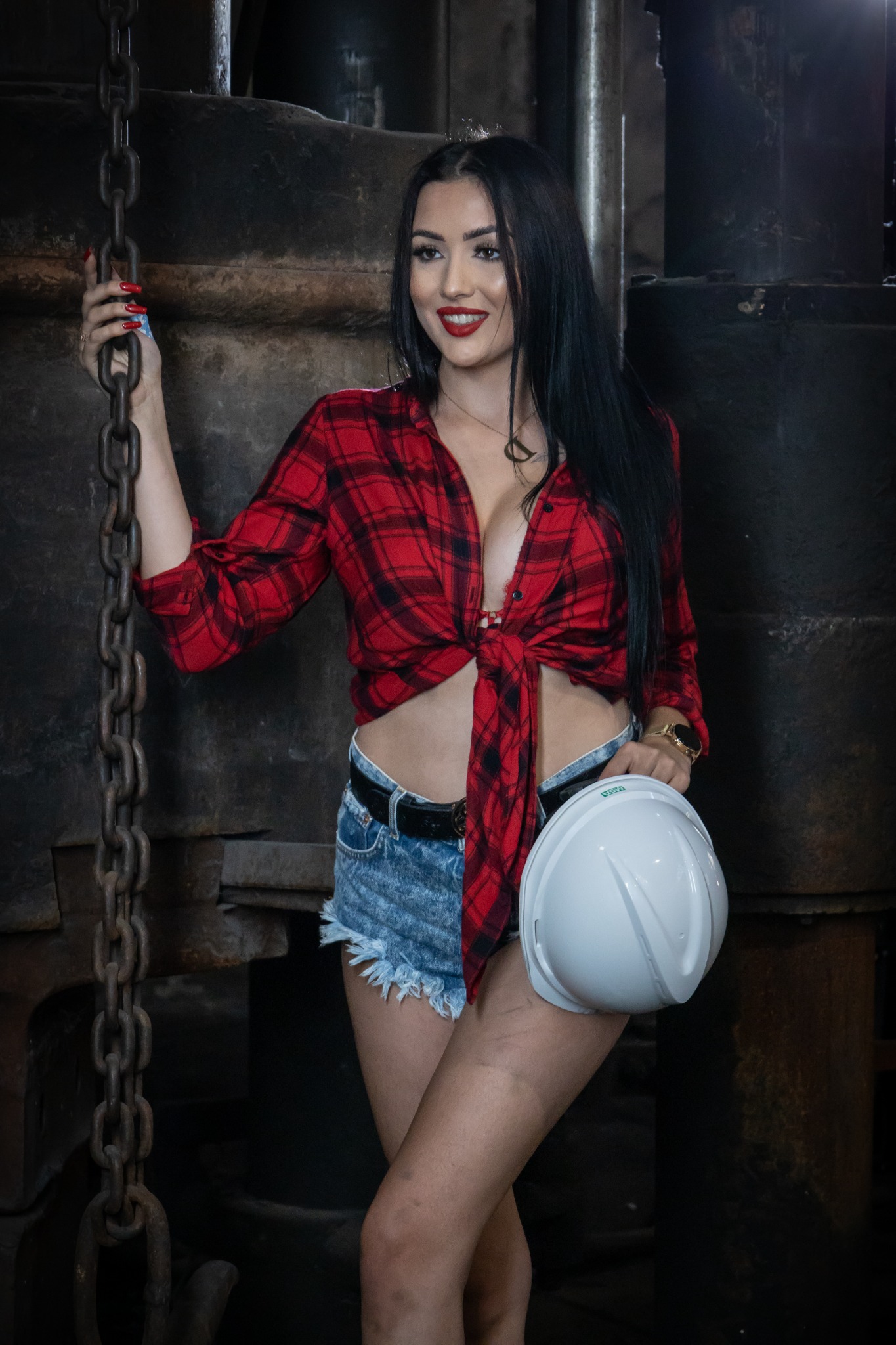 Kobieta w czerwonej koszuli w kratę, dżinsowych szortach i kasku ochronnym pozuje przy metalowej konstrukcji.