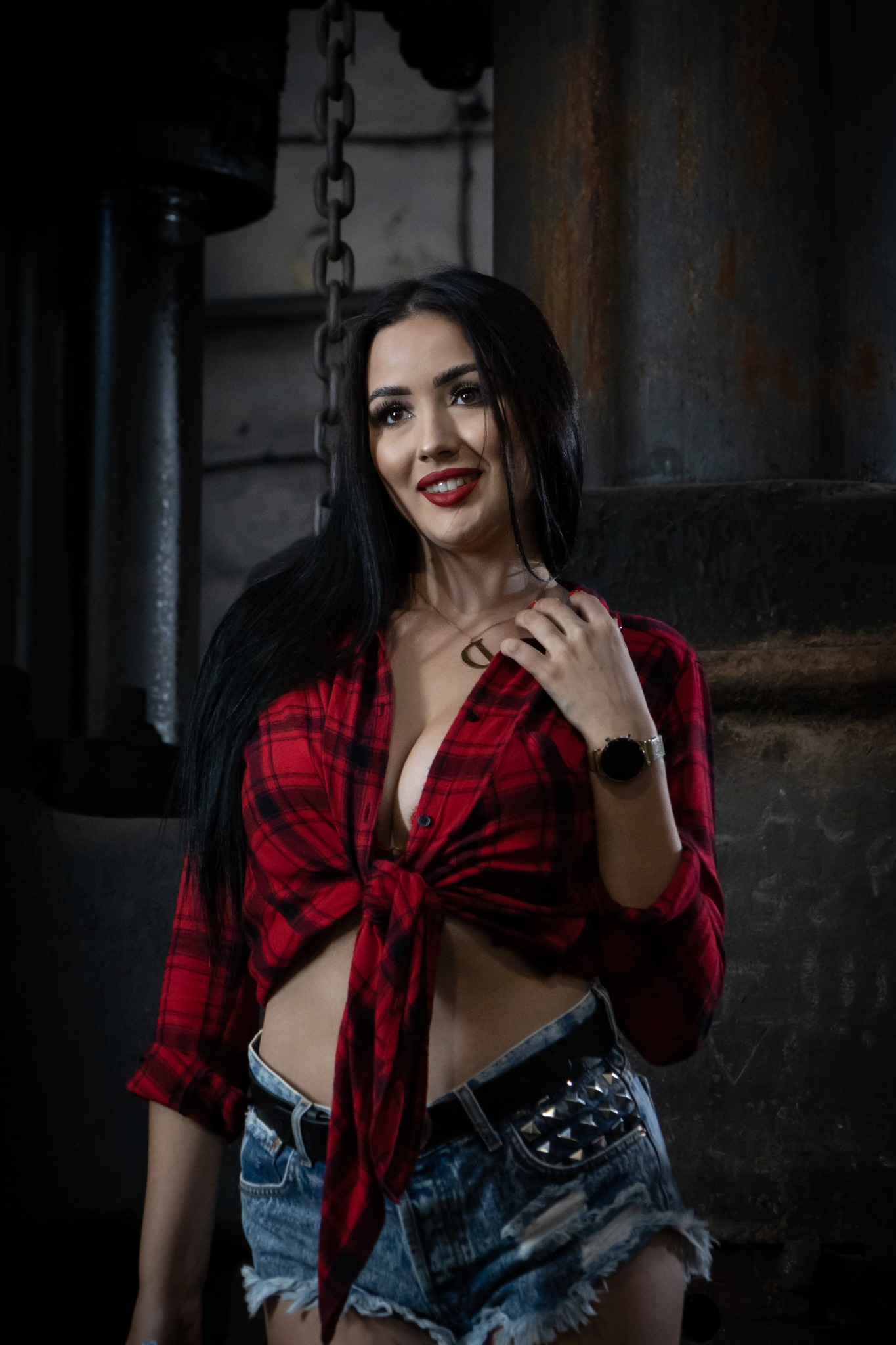 Portret kobiety w czerwonej koszuli w kratę, dżinsowych szortach z ćwiekami i złotym zegarkiem, pozującej na tle surowego, industrialnego wnętrza z łańcuchami i metalowymi elementami.