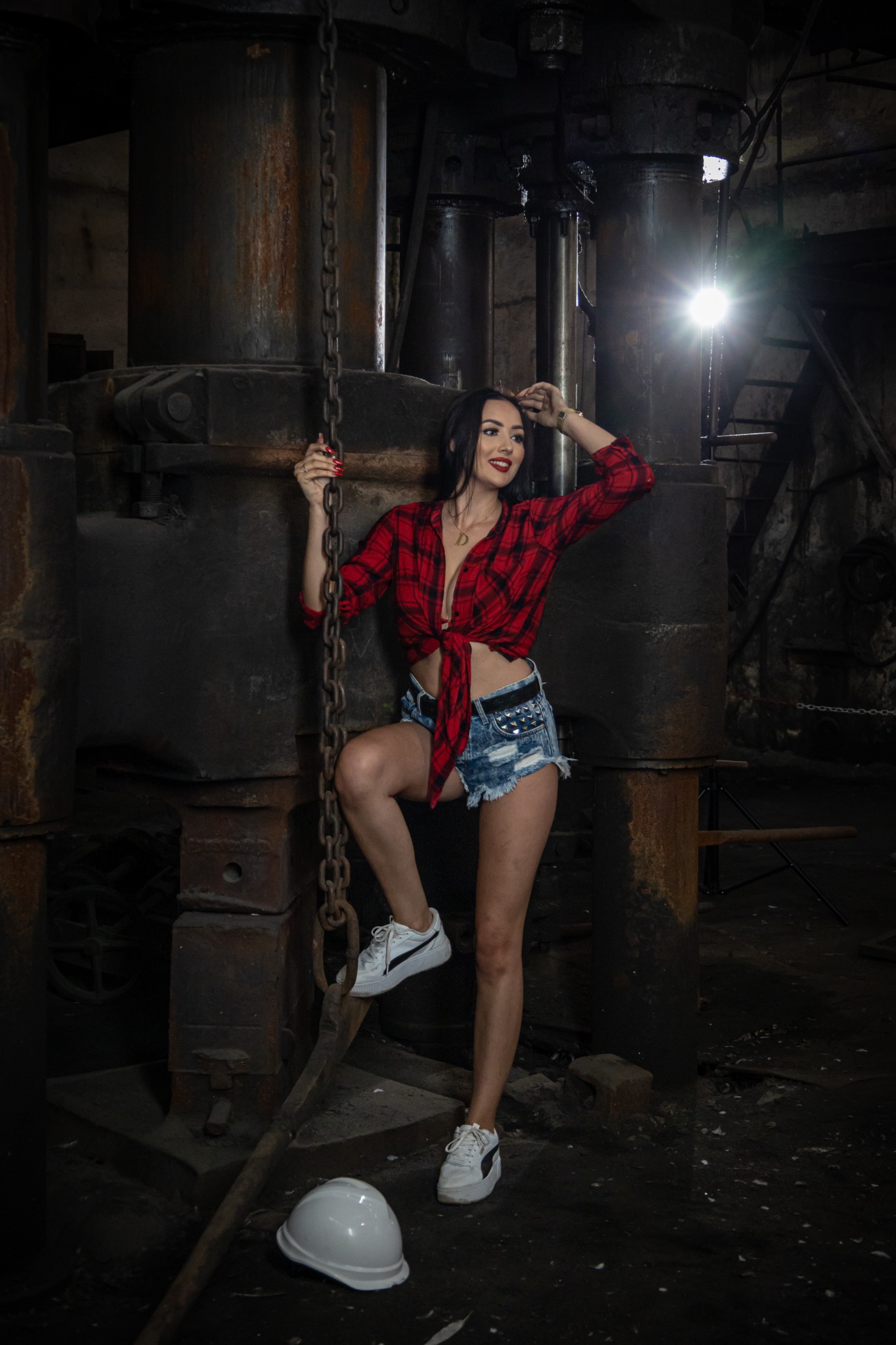 Kobieta w czerwonej koszuli w kratę i dżinsowych szortach pozuje w industrialnym otoczeniu z metalową konstrukcją i łańcuchem, obok leży biały kask ochronny.