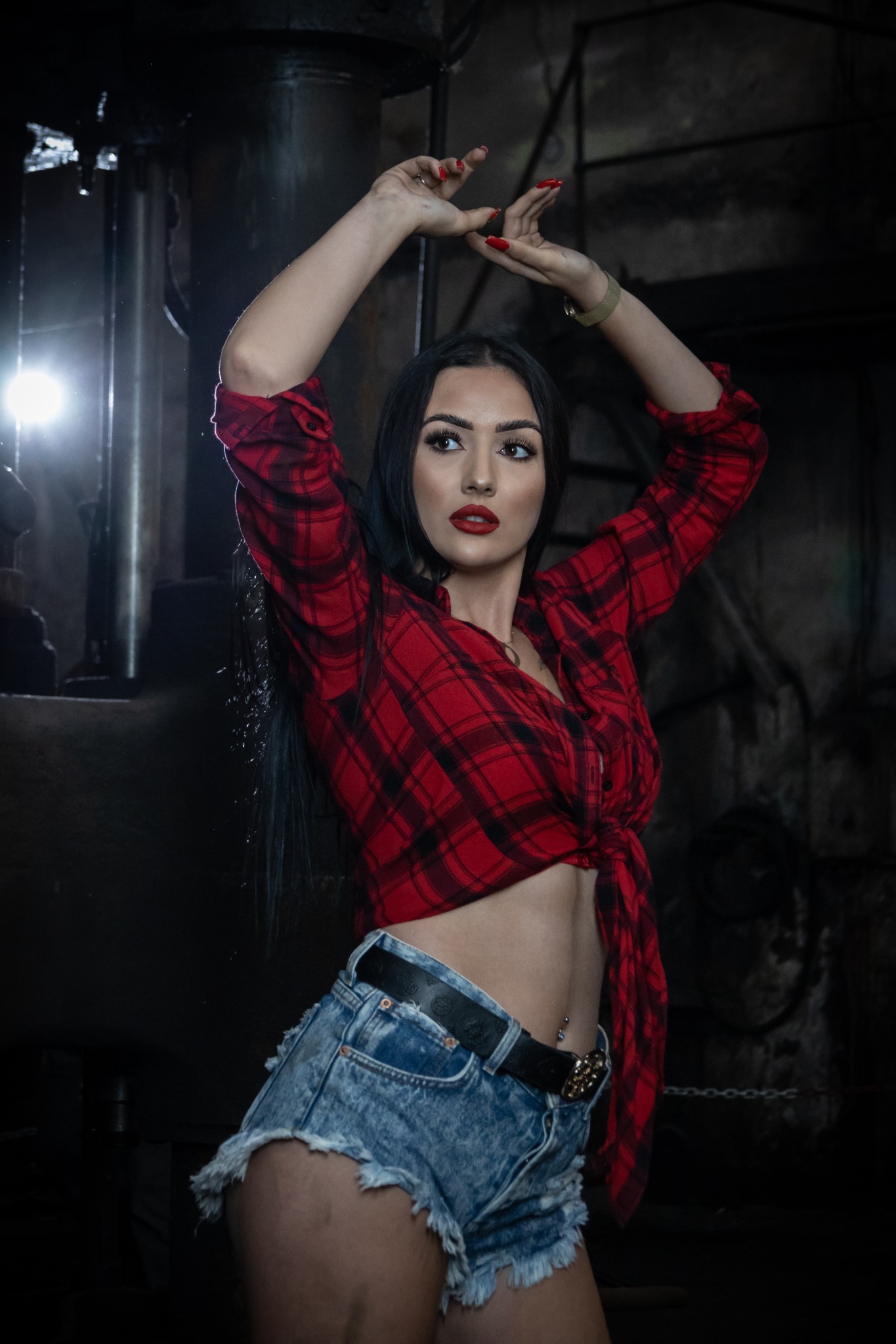 Kobieta w czerwonej koszuli w kratę i dżinsowych szortach pozuje na tle przemysłowego wnętrza z ciężkimi maszynami.