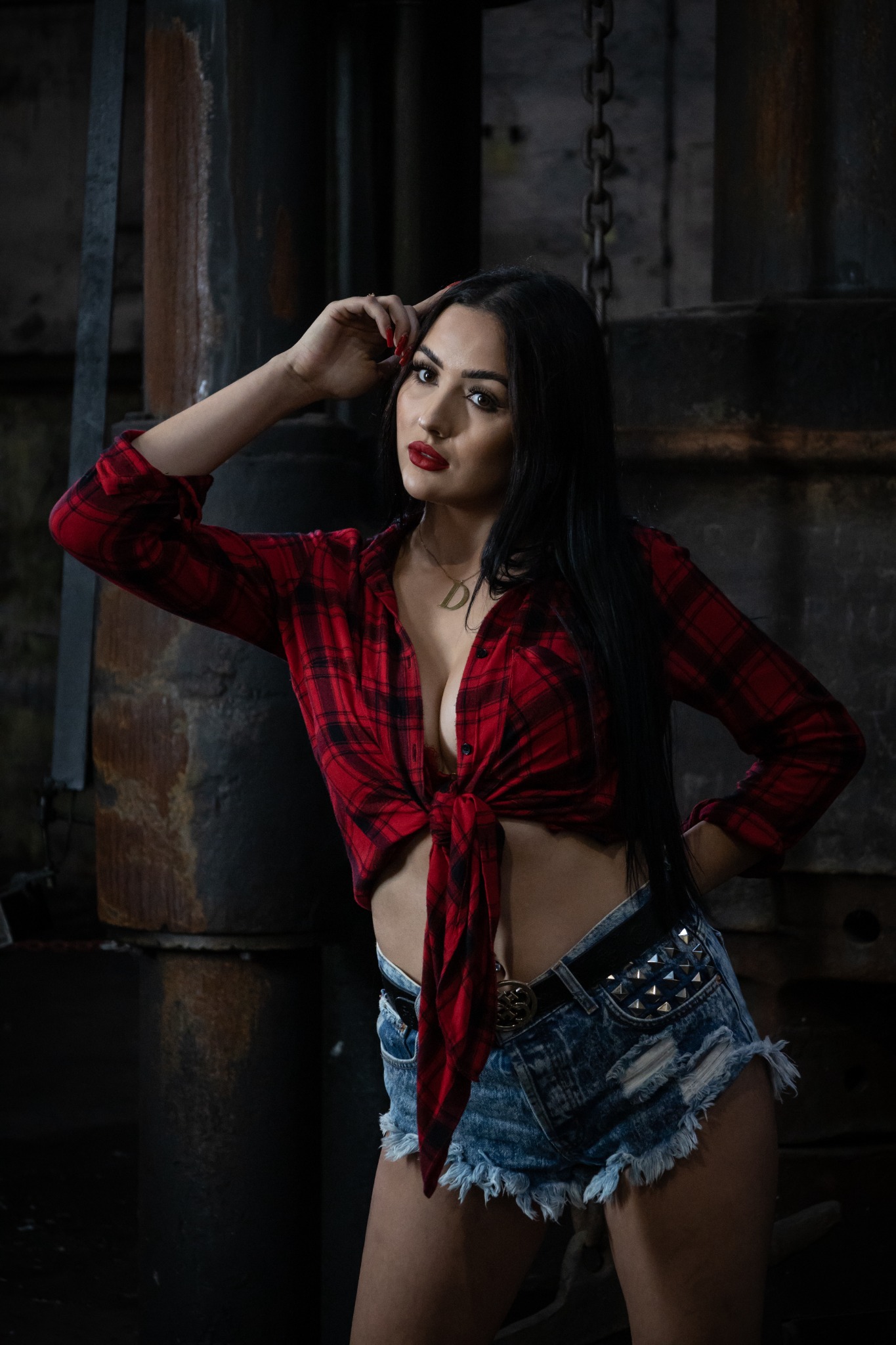 Kobieta w czerwonej koszuli w kratę i dżinsowych szortach z ćwiekami pozuje na tle industrialnego wnętrza z metalowymi elementami konstrukcyjnymi.