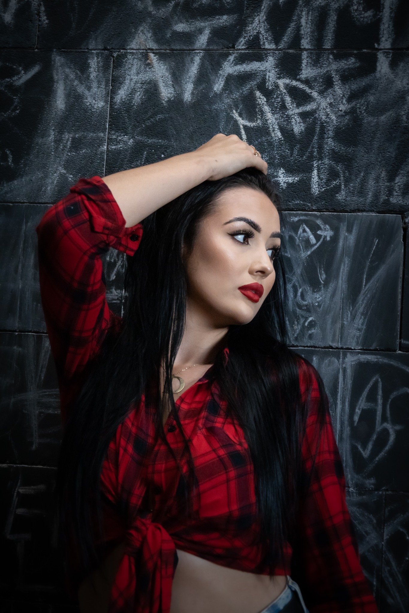 Portret kobiety z ciemnymi włosami i czerwoną koszulą w kratę, pozującej na tle ściany z graffiti.