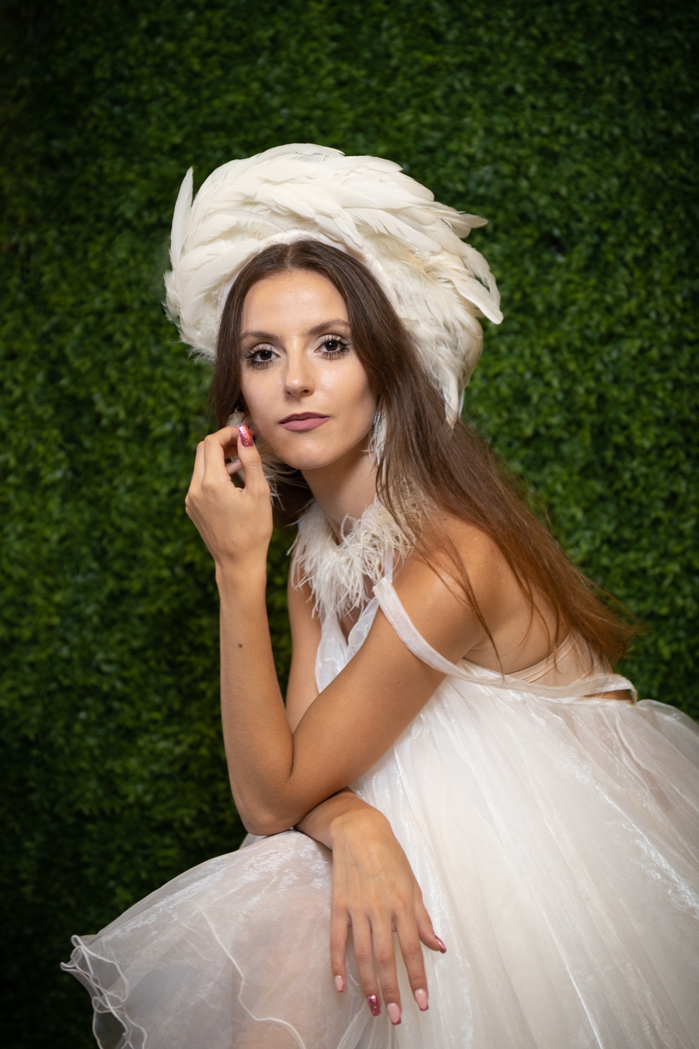 Kobieta w białej sukni i kapeluszu z piór pozuje na tle zielonej ściany. Uwagę przyciągają błyszczące paznokcie i delikatny makijaż.