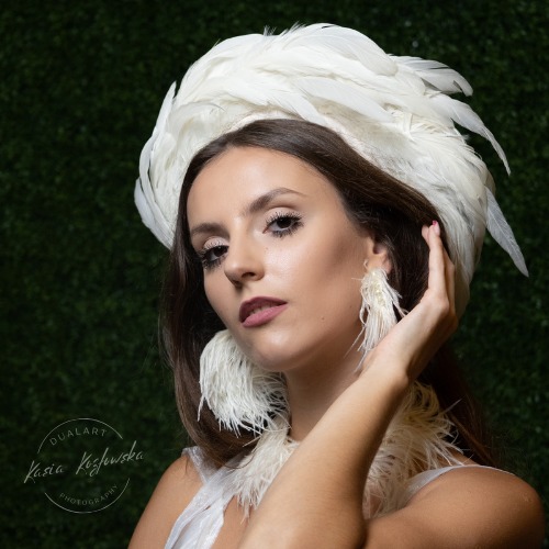 Portret kobiety w białej kreacji z piórami, ozdobnym nakryciu głowy i kolczykami na tle zielonej ściany.