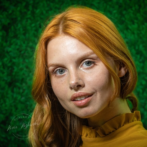 Portret rudej kobiety z piegami na tle zielonej ściany z bluszczu, w musztardowej bluzce z falbankami przy szyi, delikatny uśmiech, naturalny makijaż.