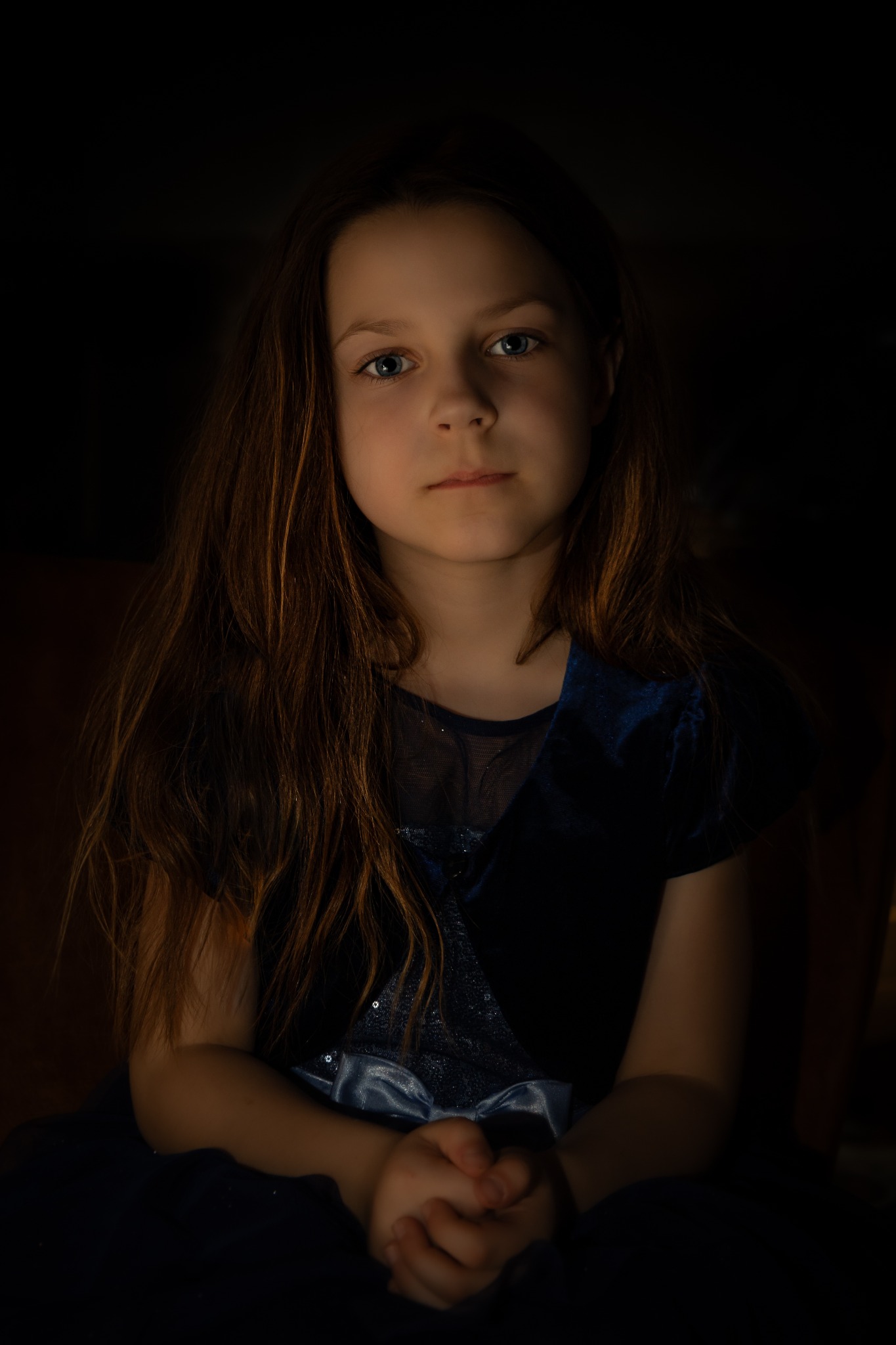 Portret dziewczynki o niebieskich oczach w ciemnoniebieskiej sukience, dłonie splecione na kolanach, oświetlona z przodu, ciemne tło.