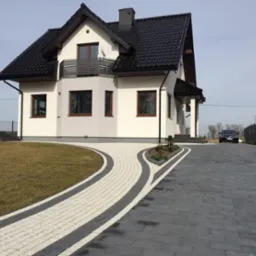 Nowoczesny dom jednorodzinny z podjazdem wyłożonym jasnoszarą kostką z ciemnoszarymi obrzeżami, prowadzącym do ciemnoszarej kostki brukowej, widoczny samochód osobowy w tle.