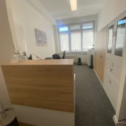 Jasne biuro kancelarii z recepcją o drewnianym blacie, szarą wykładziną i szafami na dokumenty. W tle okno z roletami i dekoracje ścienne.