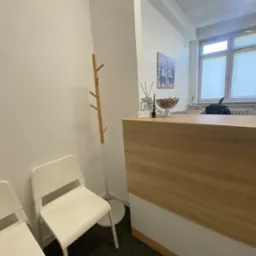 Jasne wnętrze kancelarii prawnej: białe krzesła dla klientów, drewniana lada recepcyjna z dekoracjami, w tle okno i grafika na ścianie.