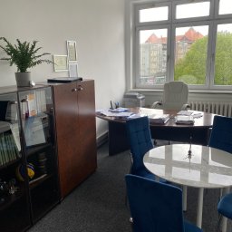 Kancelaria Radcy Prawnego Łukasz Rykowski - Wnętrze biura z biurkiem, krzesłami i regałem. Na regale książki, dyplomy i piłka do siatkówki. Za oknem widok na budynki. Neutralna kolorystyka wnętrza.