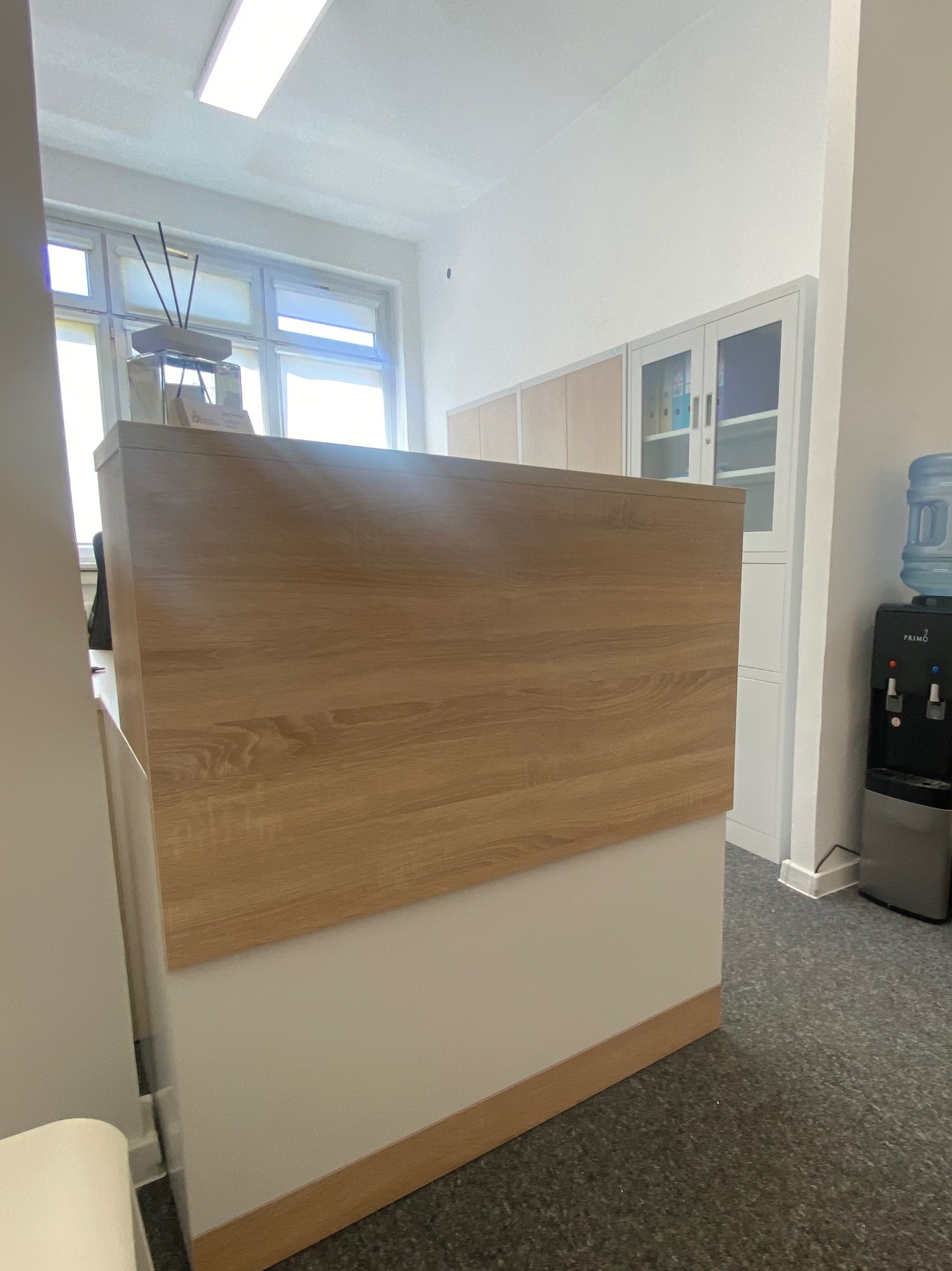Recepcja z drewnianym blatem i białym frontem, widok z boku, w tle szafy biurowe i dystrybutor wody. Jasne, neutralne oświetlenie biurowe.