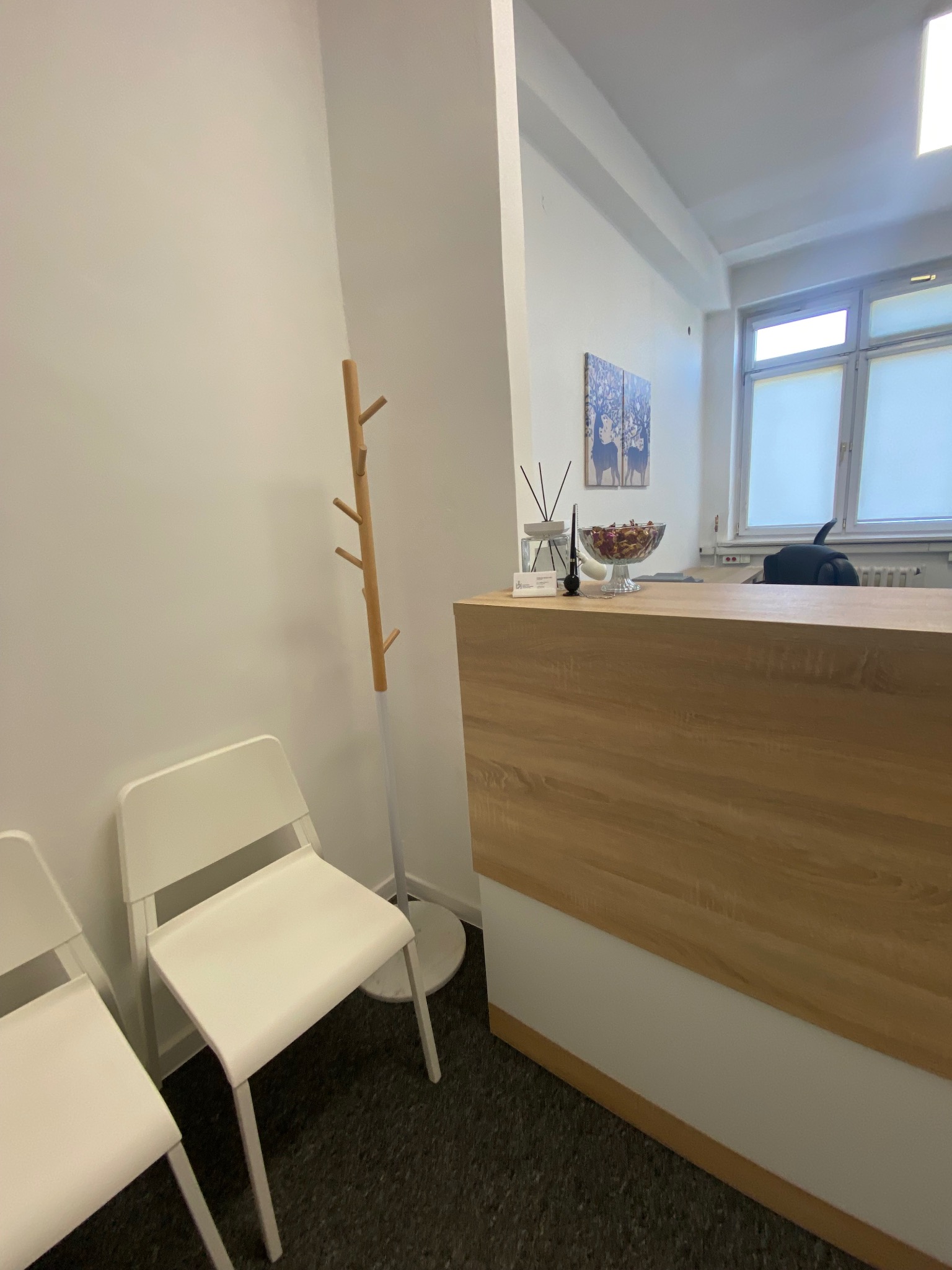 Jasne wnętrze kancelarii prawnej: białe krzesła dla klientów, drewniana lada recepcyjna z dekoracjami, w tle okno i grafika na ścianie.