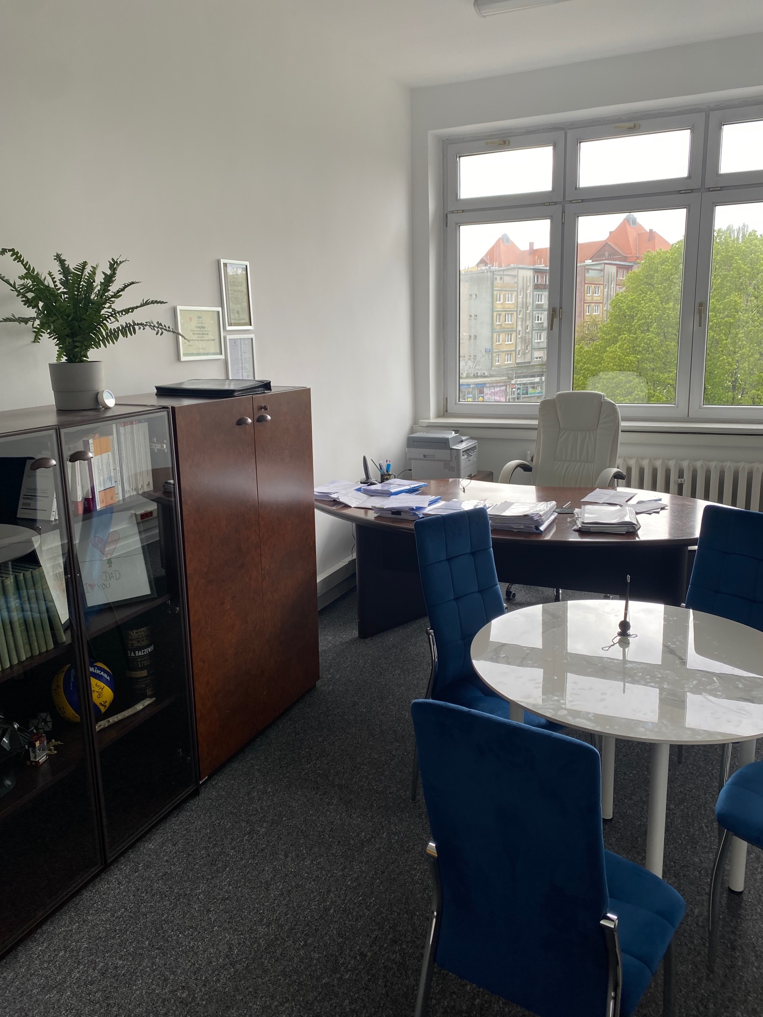 Wnętrze biura z biurkiem, krzesłami i regałem. Na regale książki, dyplomy i piłka do siatkówki. Za oknem widok na budynki. Neutralna kolorystyka wnętrza.
