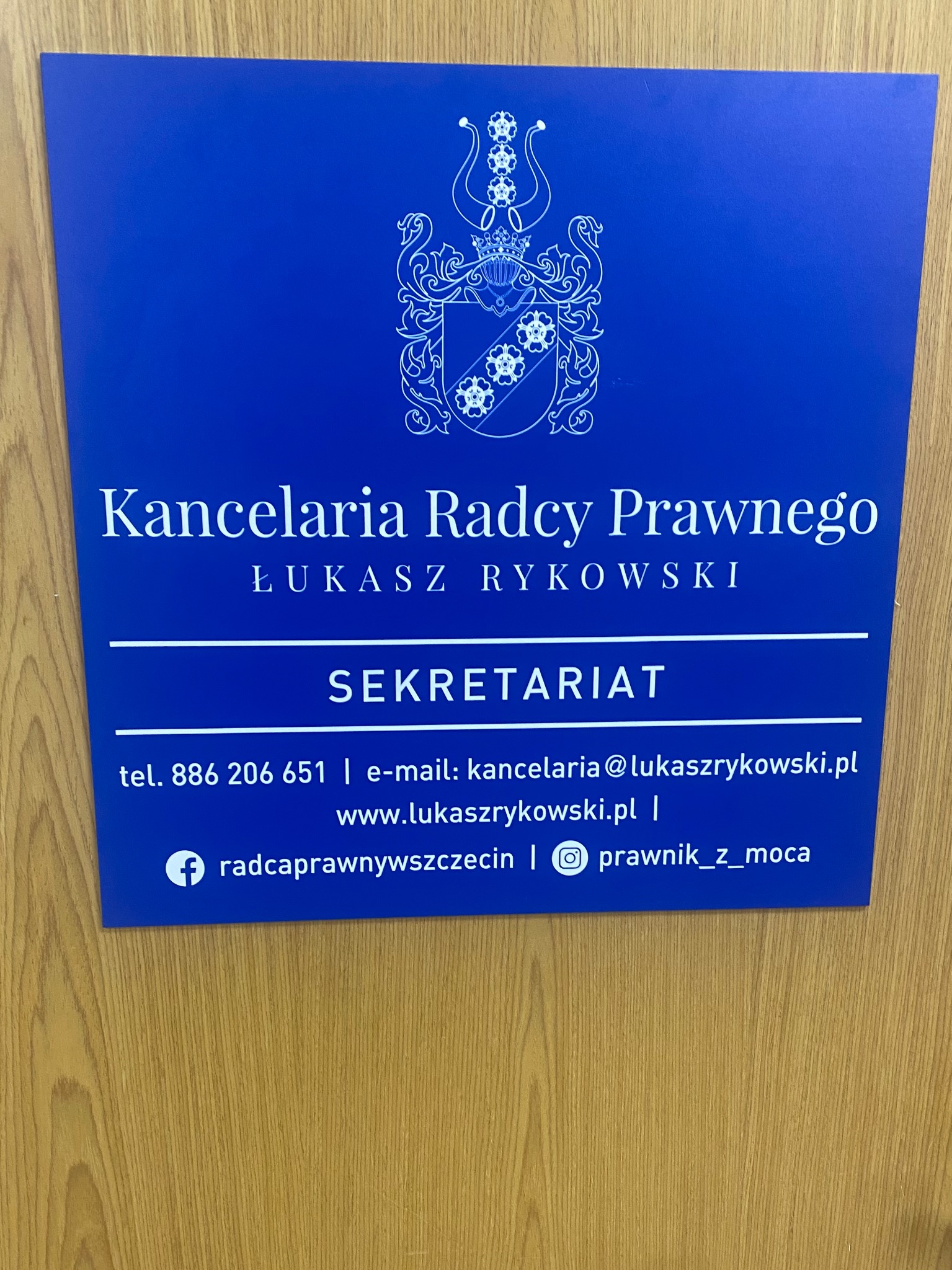 Niebieska tabliczka informacyjna z herbem i danymi kontaktowymi Kancelarii Radcy Prawnego Łukasza Rykowskiego, umieszczona na drewnianym tle.