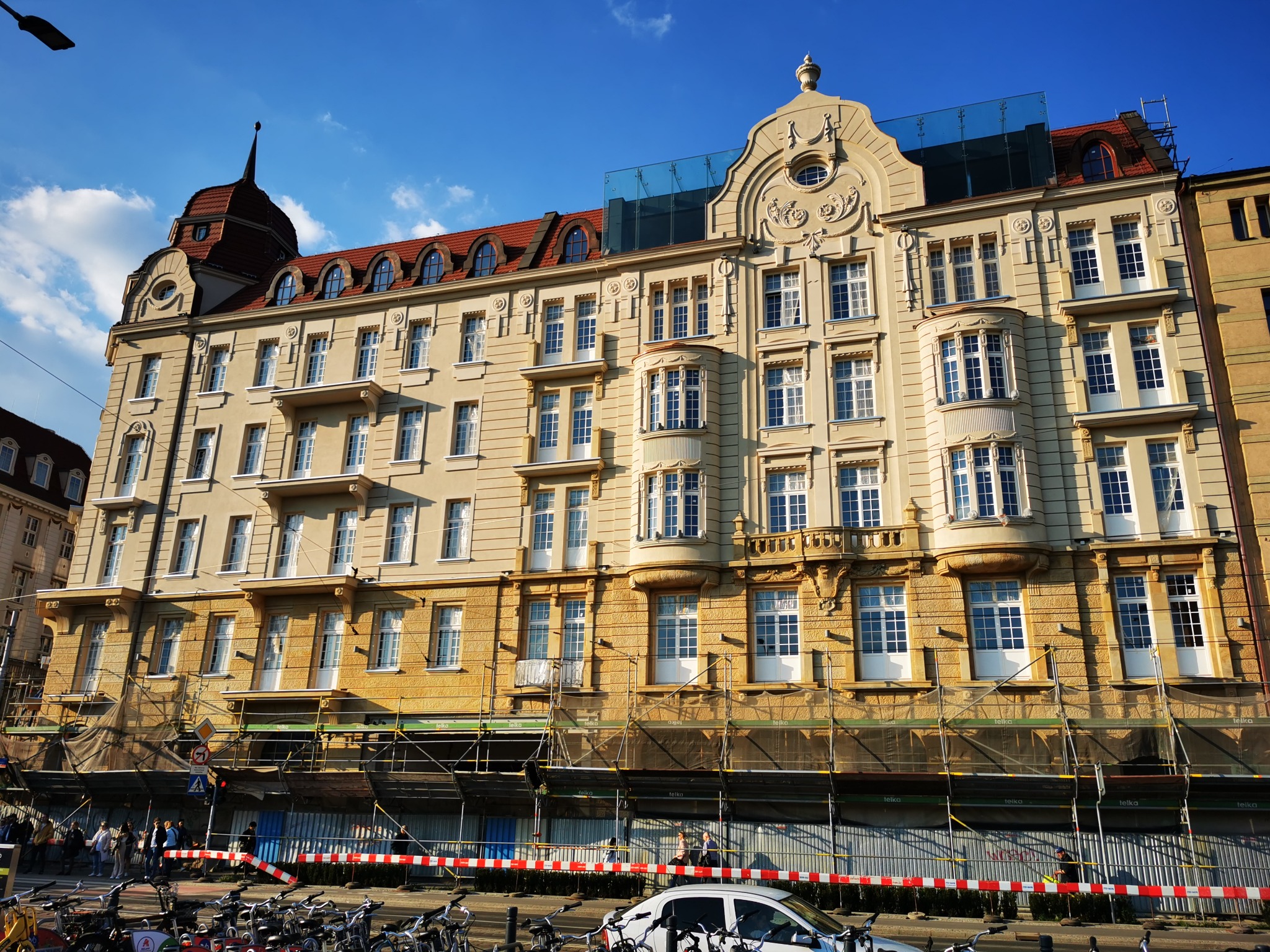 Elewacja Hotelu Grand we Wrocławiu w trakcie renowacji, z widocznymi rusztowaniami i siatkami ochronnymi na dolnych kondygnacjach, rowerami zaparkowanymi przed budynkiem i fragmentem chodnika...