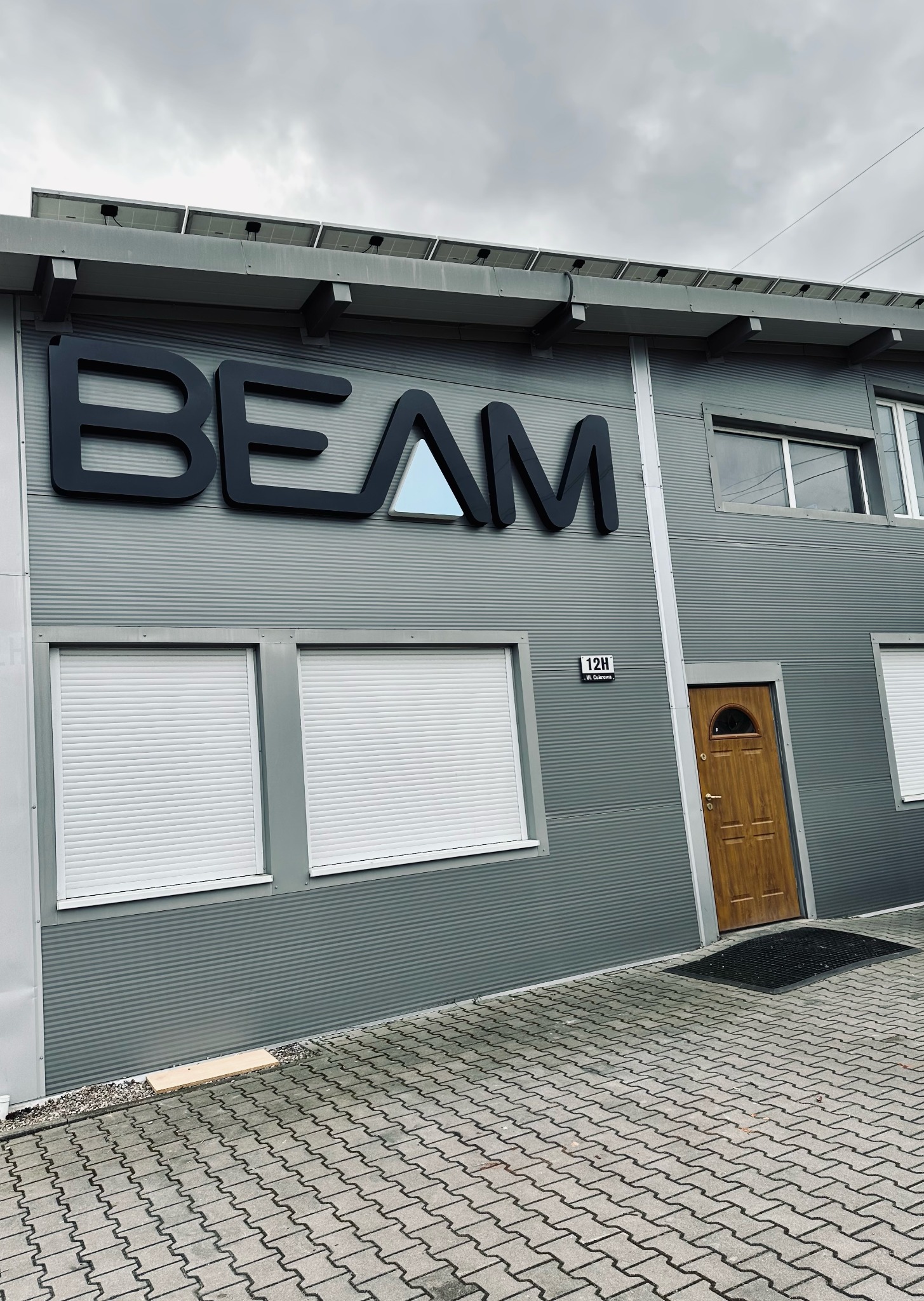 Szary budynek biurowy z napisem 'BEAM' i panelami słonecznymi na dachu, widziany pod kątem.