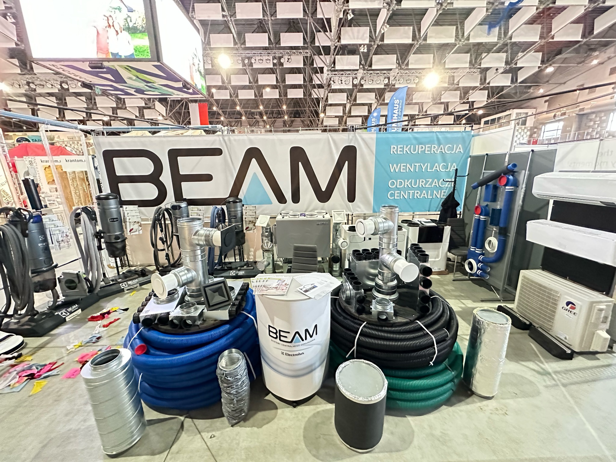 Ekspozycja na targach z systemami wentylacyjnymi i odkurzaczami centralnymi marki Beam i Electrolux, widoczne rury, kształtki, jednostki centralne i klimatyzator Gree na szarym tle hali.