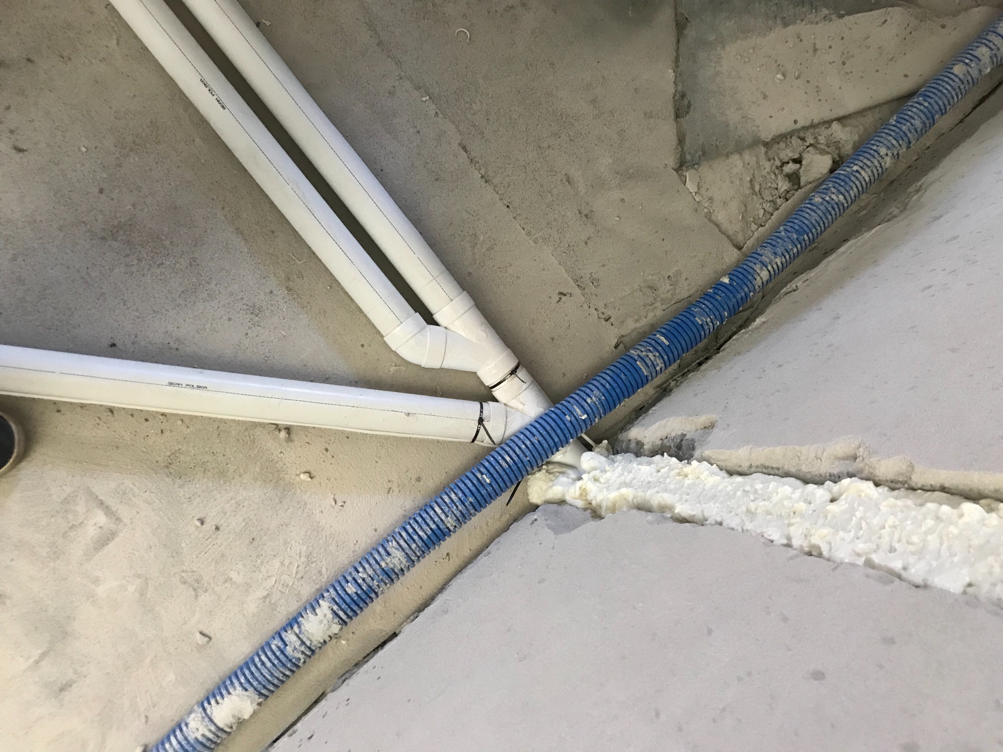 Białe rury PCV oraz niebieska rura karbowana przymocowane do sufitu, uszczelnione pianką montażową.