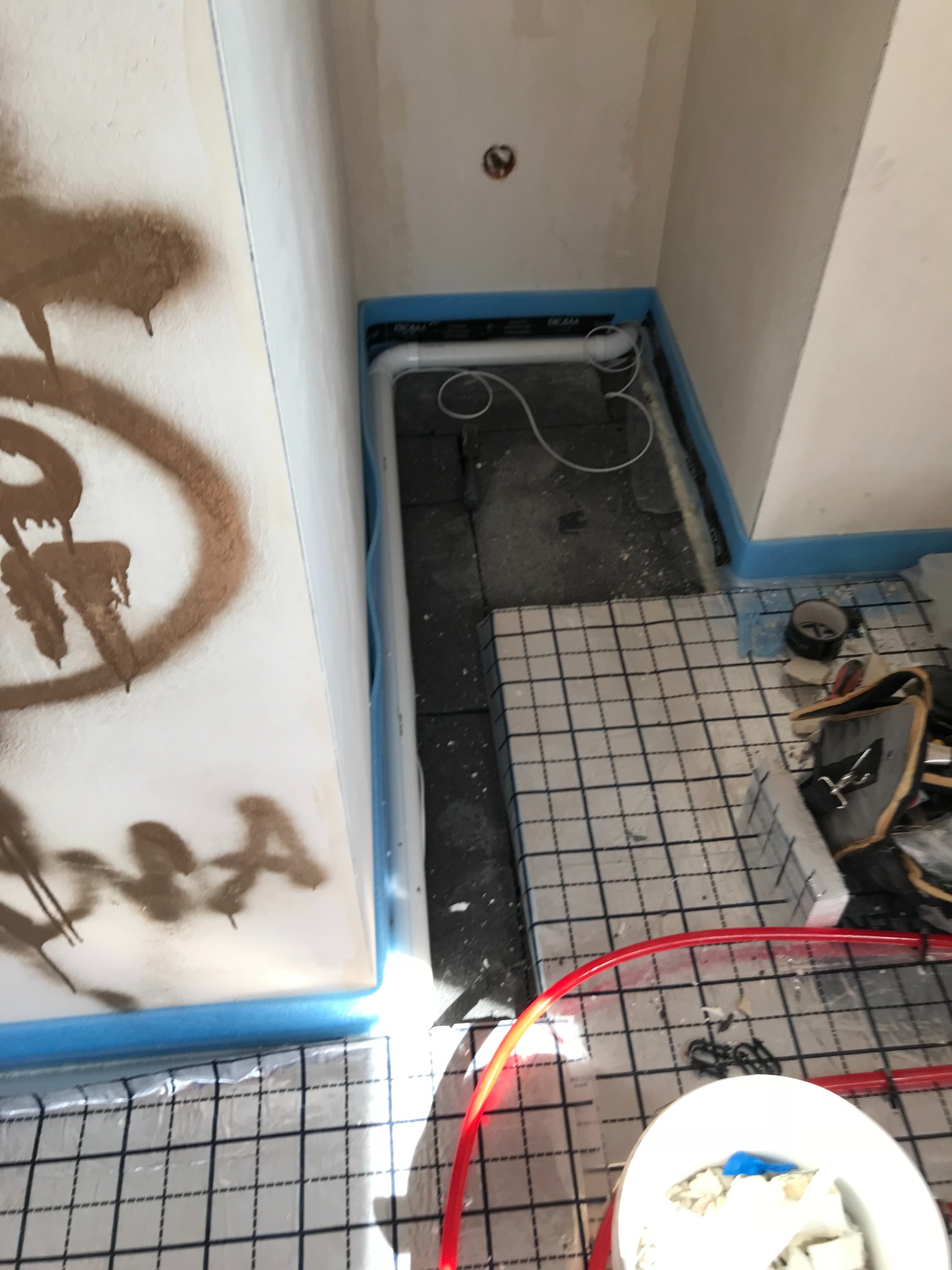 Instalacja ogrzewania podłogowego w narożniku pomieszczenia, widoczne rury grzewcze układane na izolacji z siatką, narzędzia pracy obok, ściany z widocznymi śladami prac wykończeniowych i brudnymi...