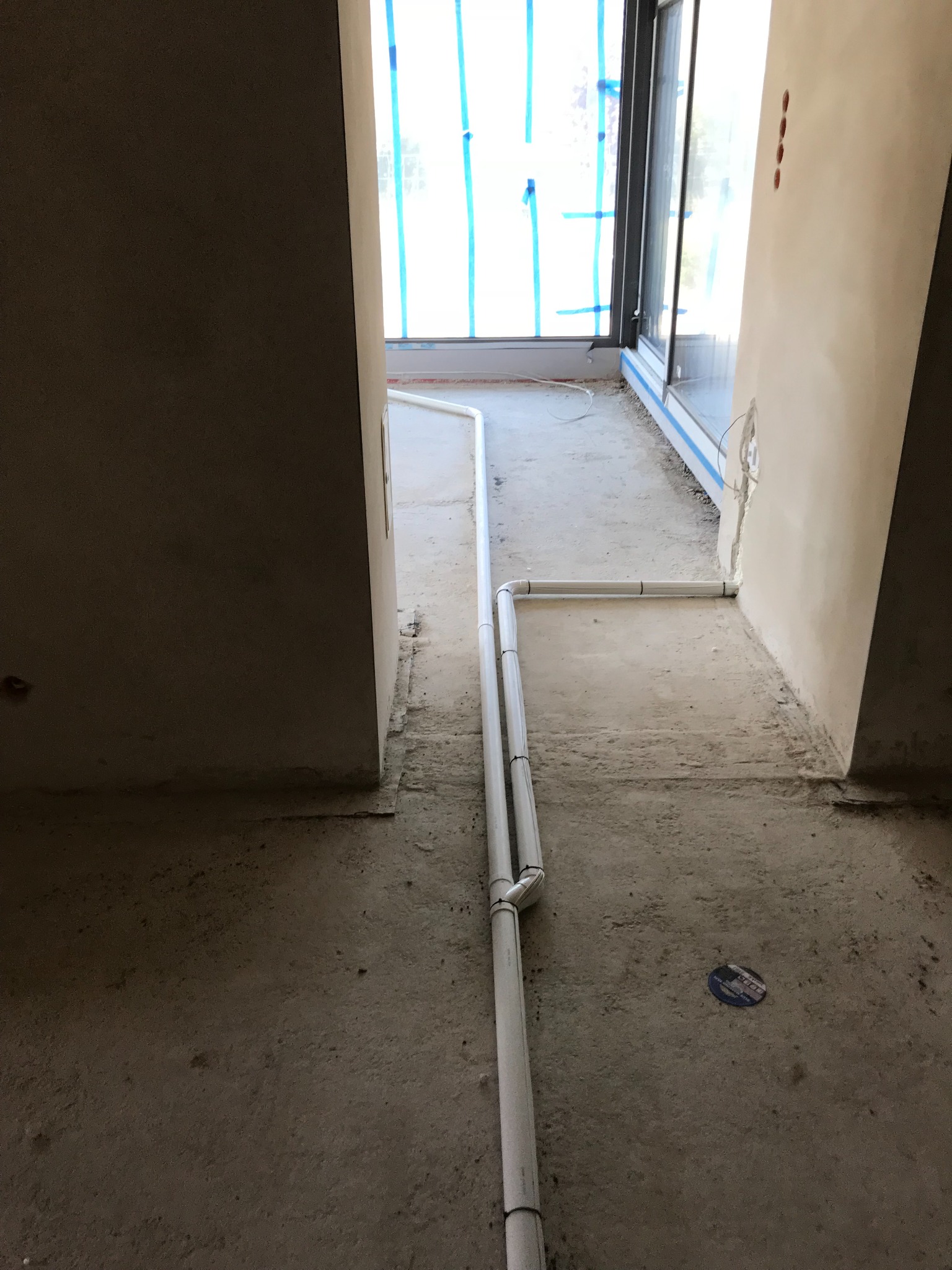 Białe rury instalacyjne ułożone na surowym betonowym podłożu w nowym budynku, widok z perspektywy niskiego kąta, ściany w stanie deweloperskim, okno zabezpieczone niebieską taśmą.