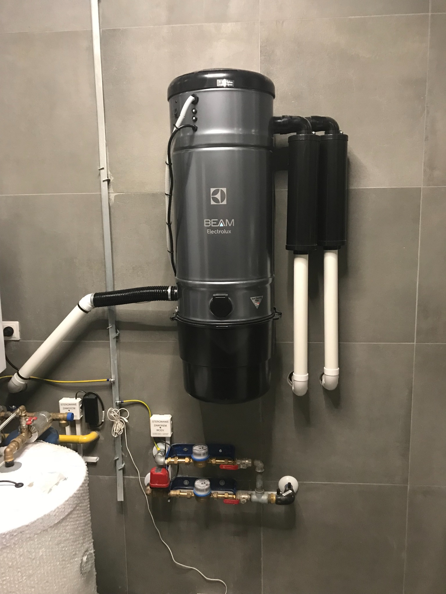 Zamontowany centralny odkurzacz BEAM Electrolux na szarej ścianie z płytkami, widoczne rury i instalacje wodne w tle.
