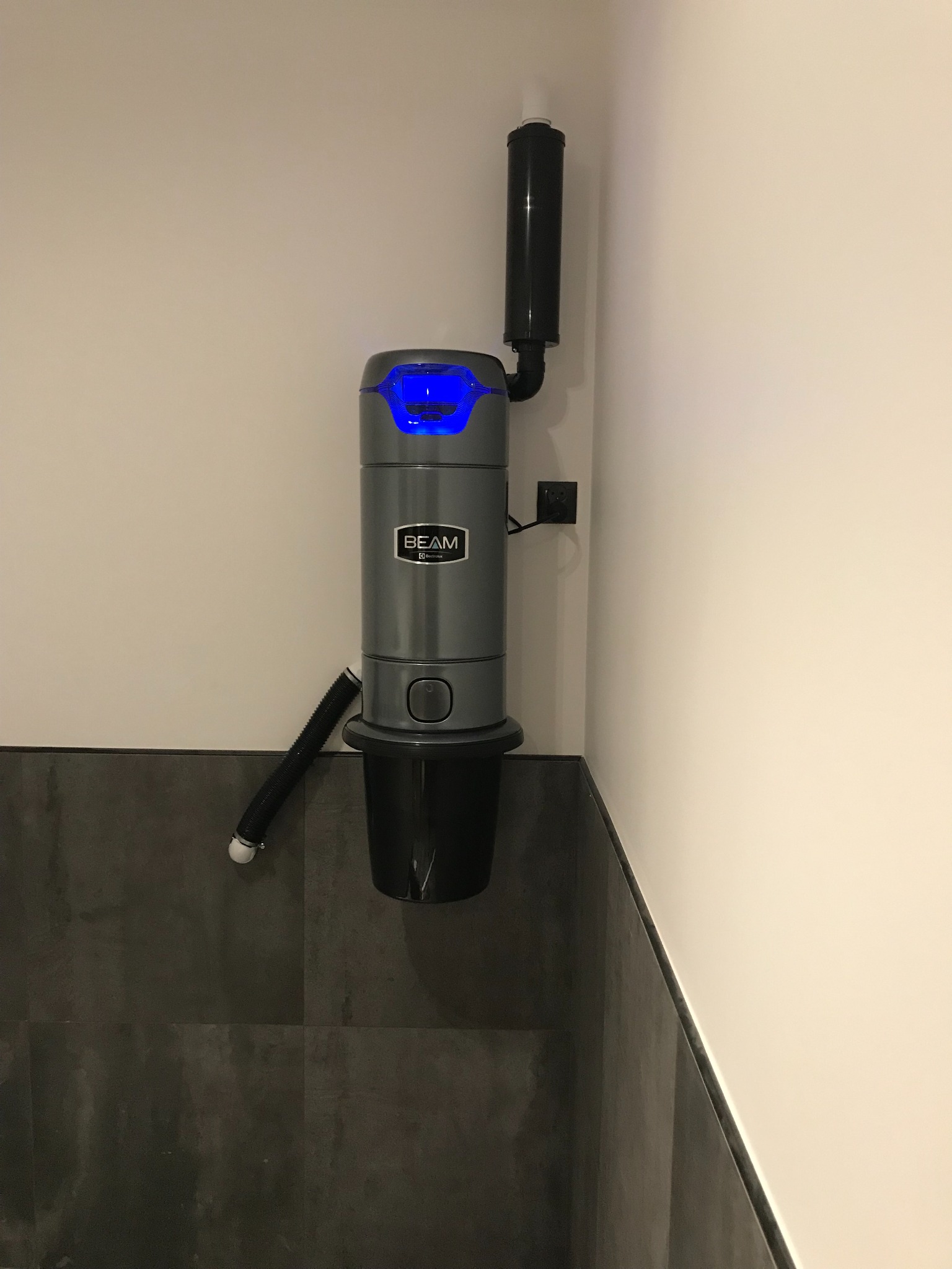 Szary odkurzacz centralny BEAM zamontowany na ścianie, z widocznym wężem i niebieskim podświetleniem, na tle szarej ściany i białego narożnika.