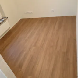 Nowo ułożona podłoga z paneli w odcieniach brązu w jasnym pokoju z białymi ścianami, widoczne gniazdka elektryczne i grzejnik.