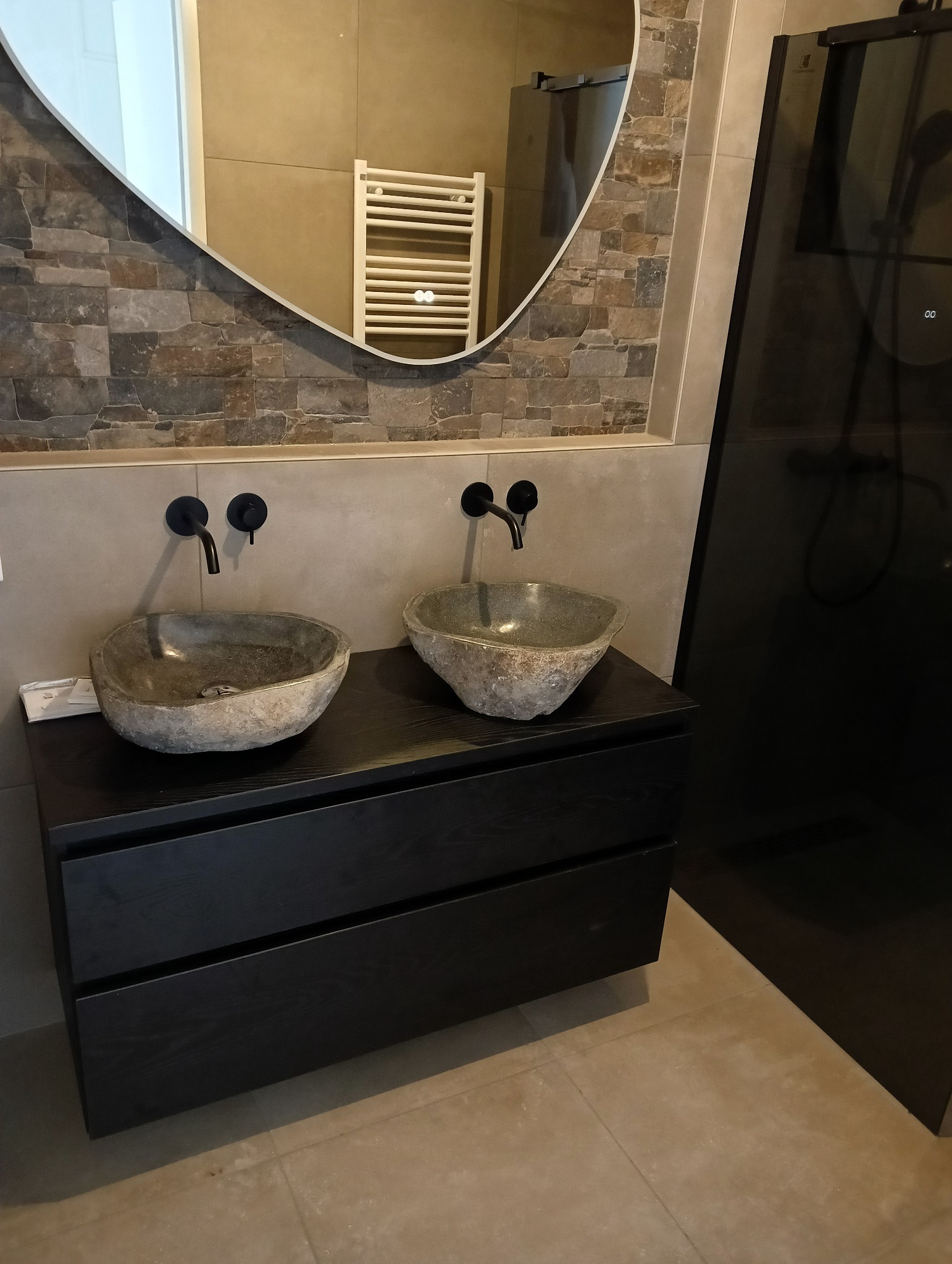 Stylowa łazienka z kamienną mozaiką, owalnym lustrem i dwiema umywalkami z naturalnego kamienia na czarnej szafce z szufladami oraz kabiną prysznicową.