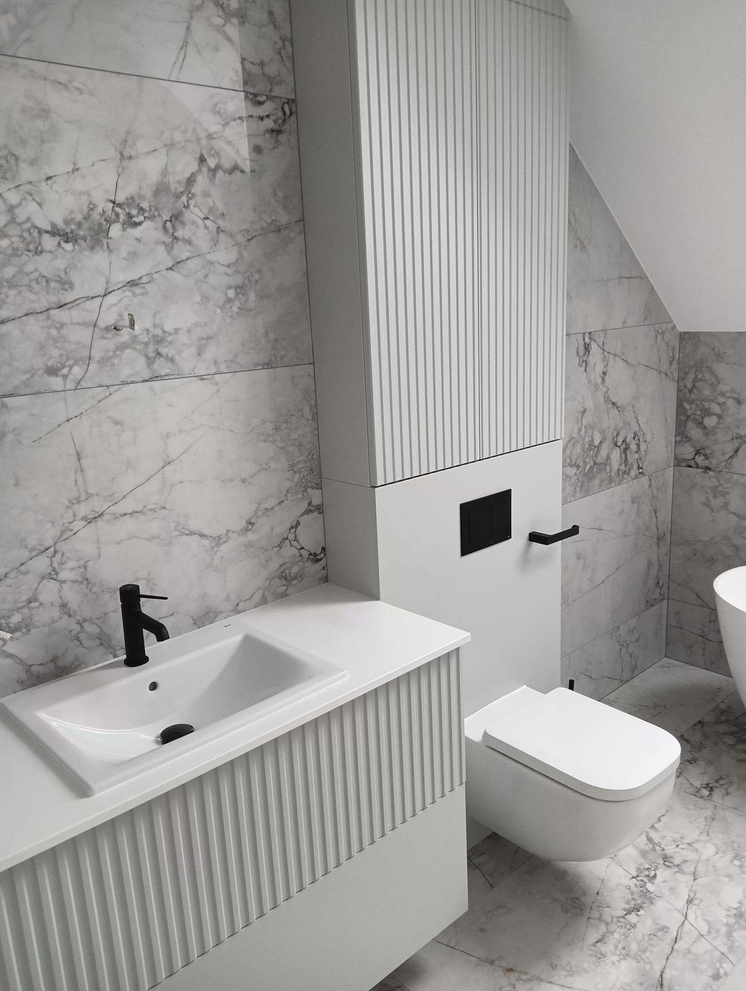 Nowoczesna łazienka w odcieniach szarości i bieli z marmurowymi płytkami, wisząca toaleta i umywalka z czarną armaturą.