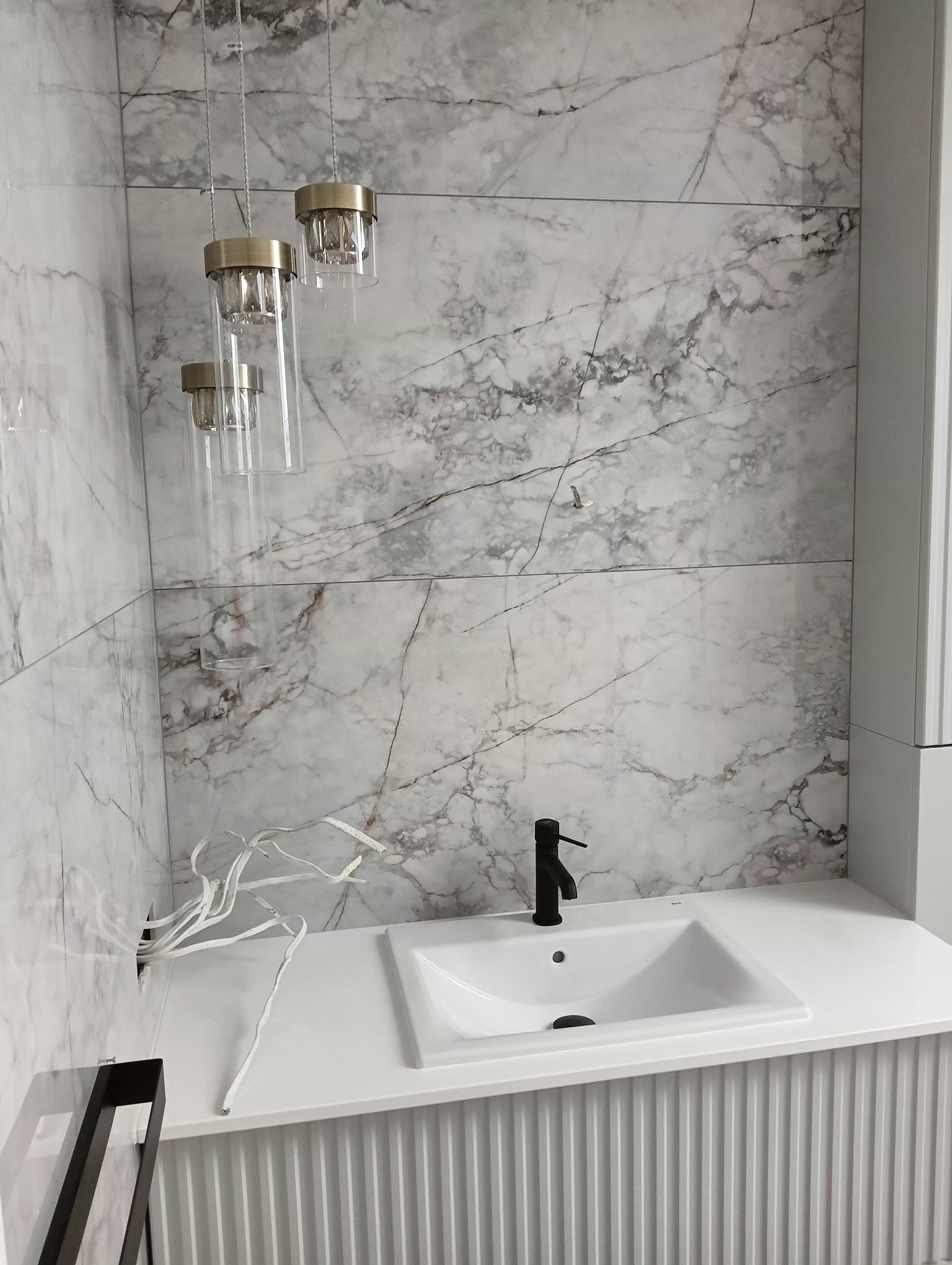 Nowoczesna łazienka z marmurowymi płytkami, czarną armaturą i wiszącymi szklanymi lampami z mosiężnymi akcentami, dekoracyjna gałązka w tle.