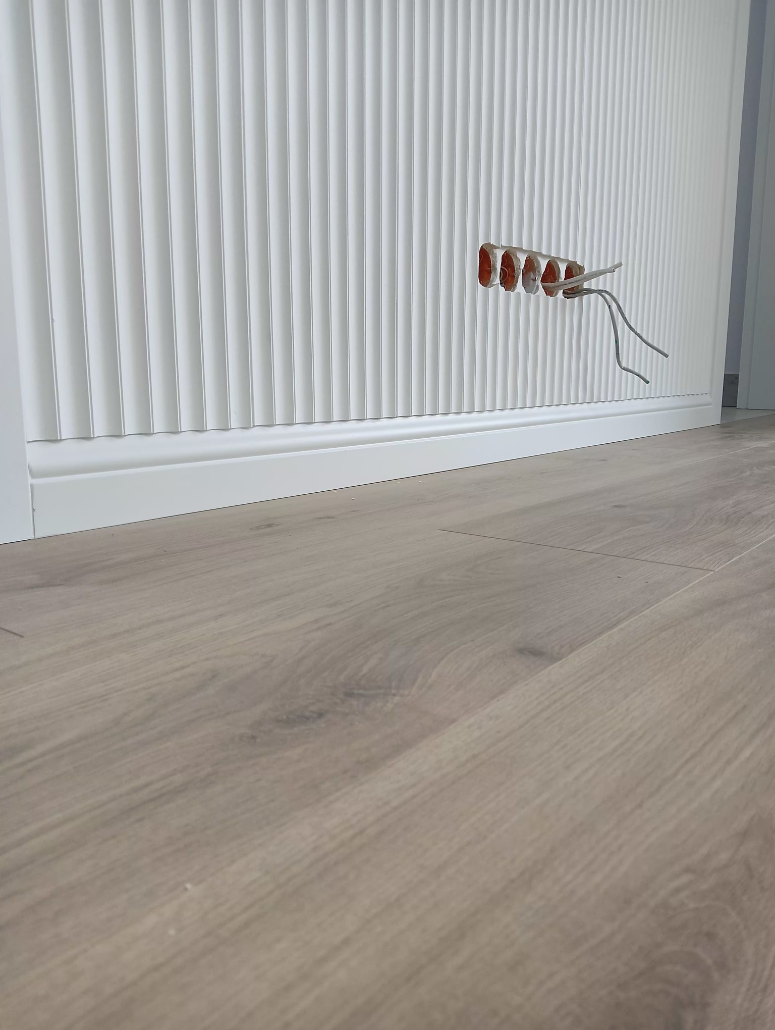 Ściana z dekoracyjnymi panelami ściennymi i listwą przypodłogową w trakcie montażu, widoczne przewody elektryczne wystające ze ściany, podłoga wyłożona panelami.