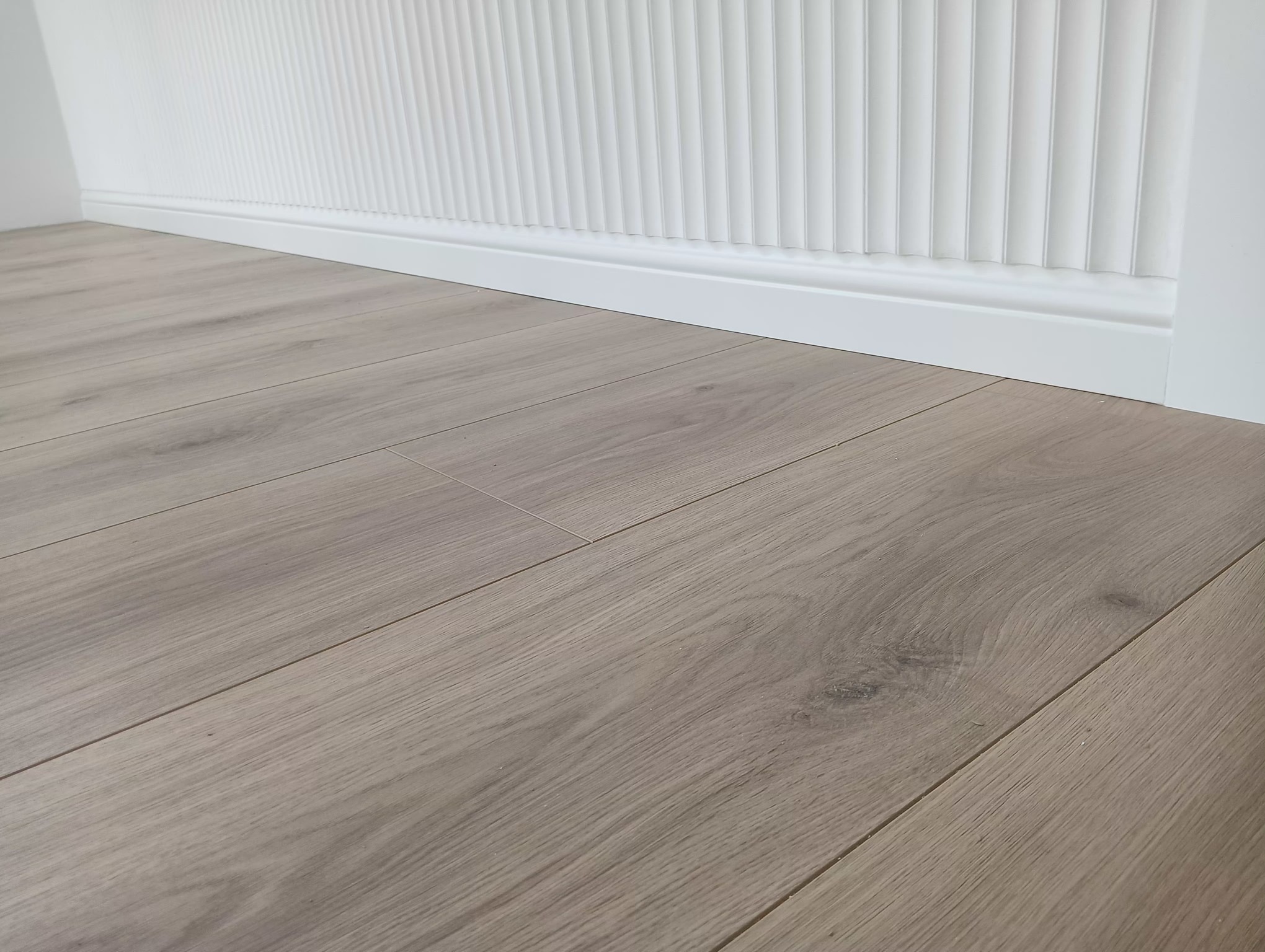 Jasne panele podłogowe ułożone wzdłuż białej listwy przypodłogowej i dekoracyjnej ściany z pionowymi żłobieniami.