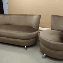 Zestaw wypoczynkowy: sofa dwuosobowa i fotel w brązowym welurze na chromowanych n&oacute;żkach, widoczne w warsztacie na tle ścian i drewnianych element&oacute;w.