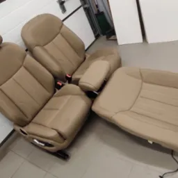 Zdemontowane, beżowe sk&oacute;rzane fotele samochodowe ułożone na posadzce garażu, widoczne elementy regulacji i mocowania.