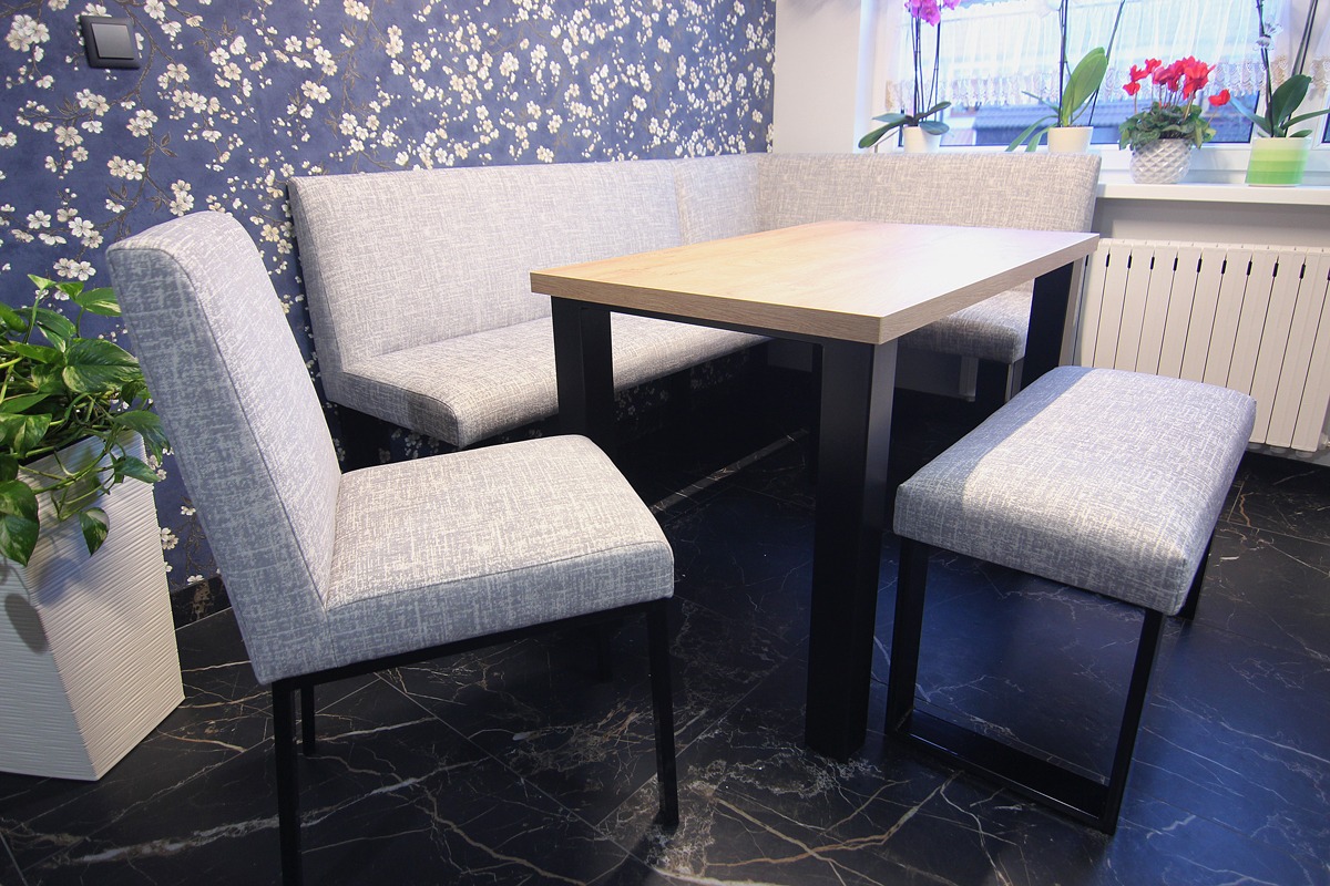 Jasnoszara tapicerowana ławka narożna z krzesłem i pufem przy stole z drewnianym blatem na tle ściany w kwiatowy wzór.