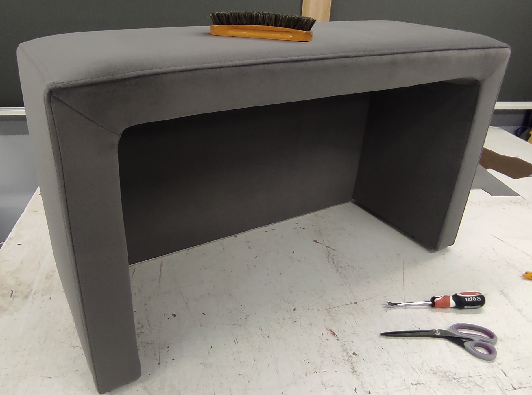Szary, tapicerowany stołek z zaokrąglonymi krawędziami, szczotka do czyszczenia tapicerki na górze, obok leżą nożyczki i śrubokręt do tapicerowania.