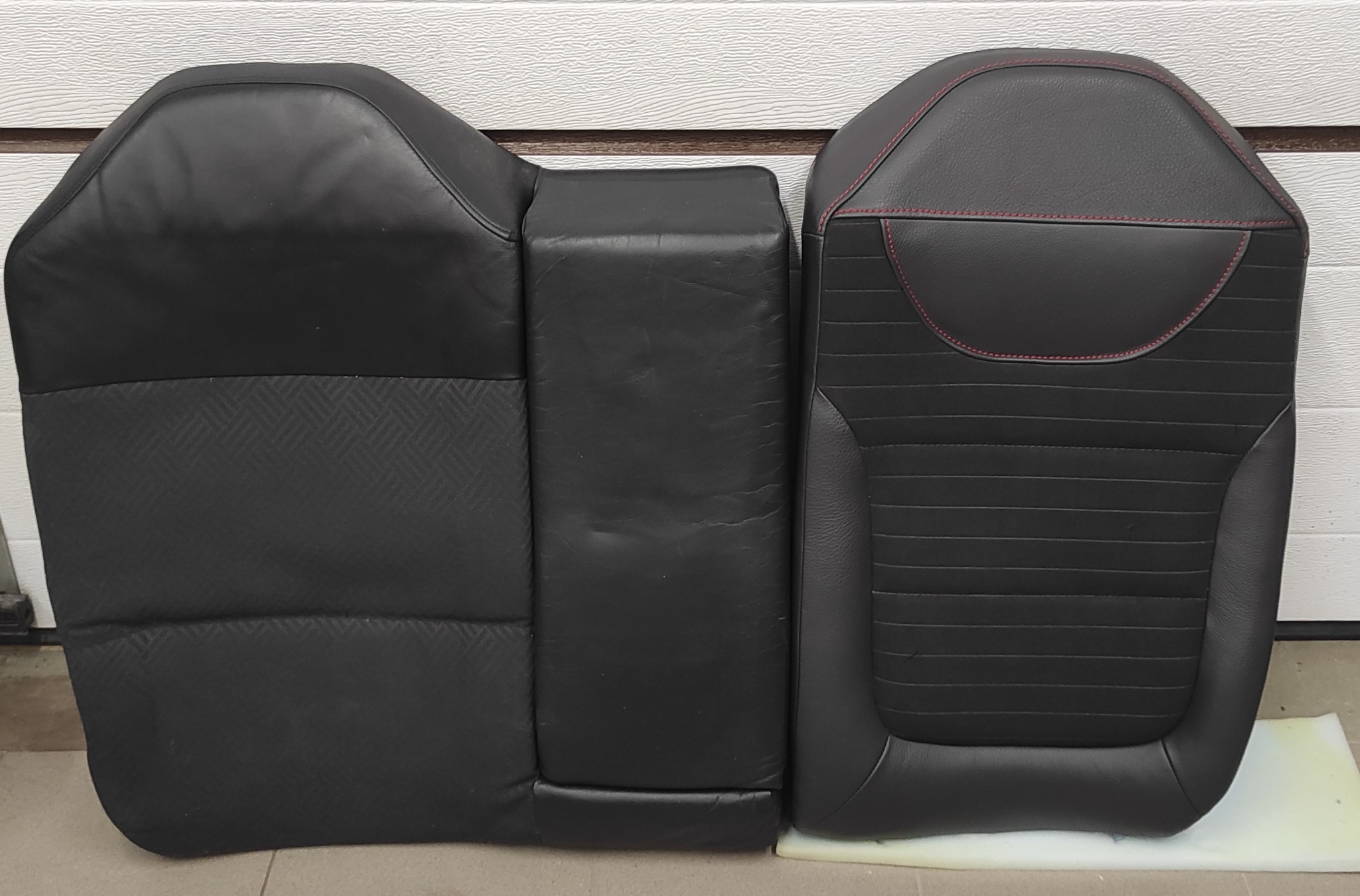 Częściowo rozebrany, czarny fotel samochodowy z widocznym fragmentem oparcia z czerwonymi przeszyciami i fragmentem siedziska o wzorzystej fakturze, leżący na podłodze warsztatu.