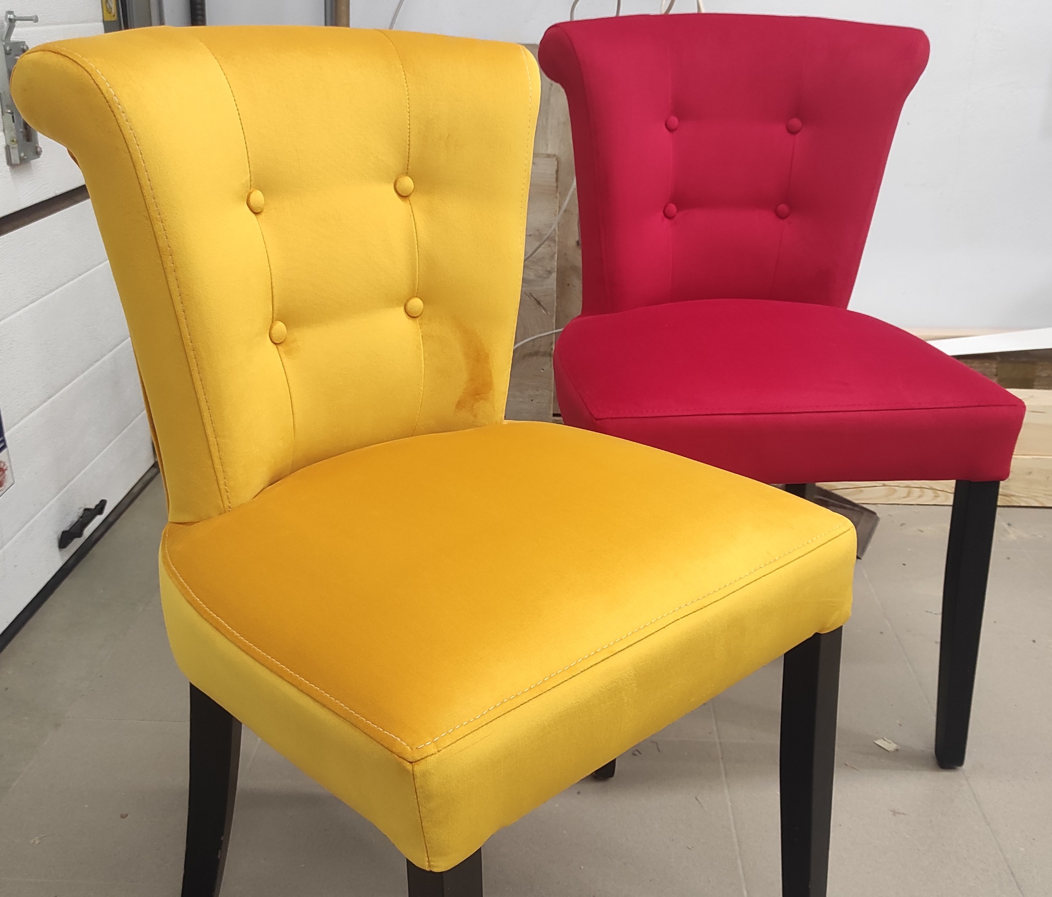 Dwa krzesła tapicerowane: jedno żółte z plamą na oparciu, drugie czerwone, oba z czarnymi nogami i guzikami na oparciu, na tle białej ściany.