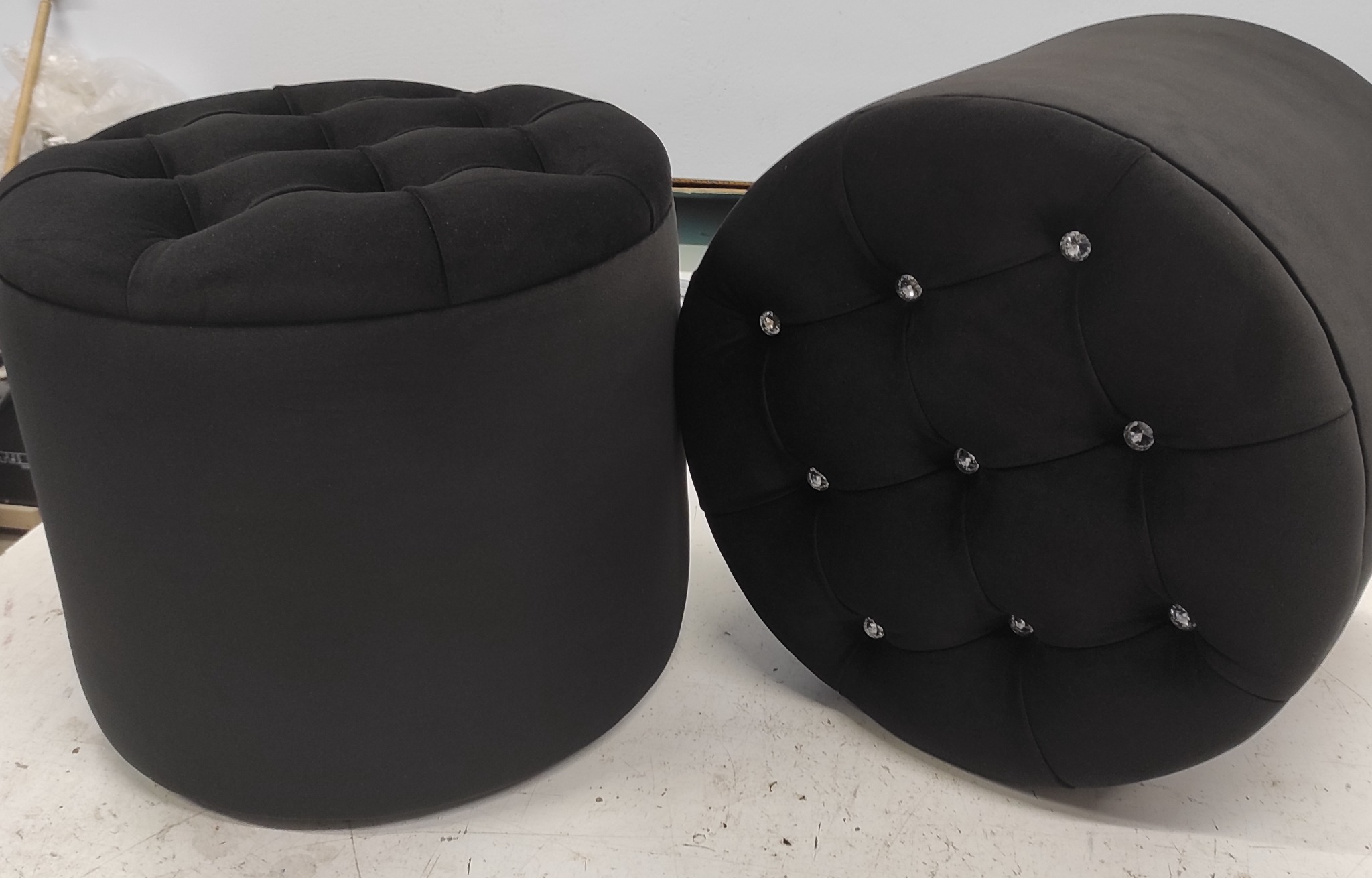 Dwa czarne, okrągłe pufy tapicerowane, jeden z guzikami, drugi z kryształkami, na jasnym tle.
