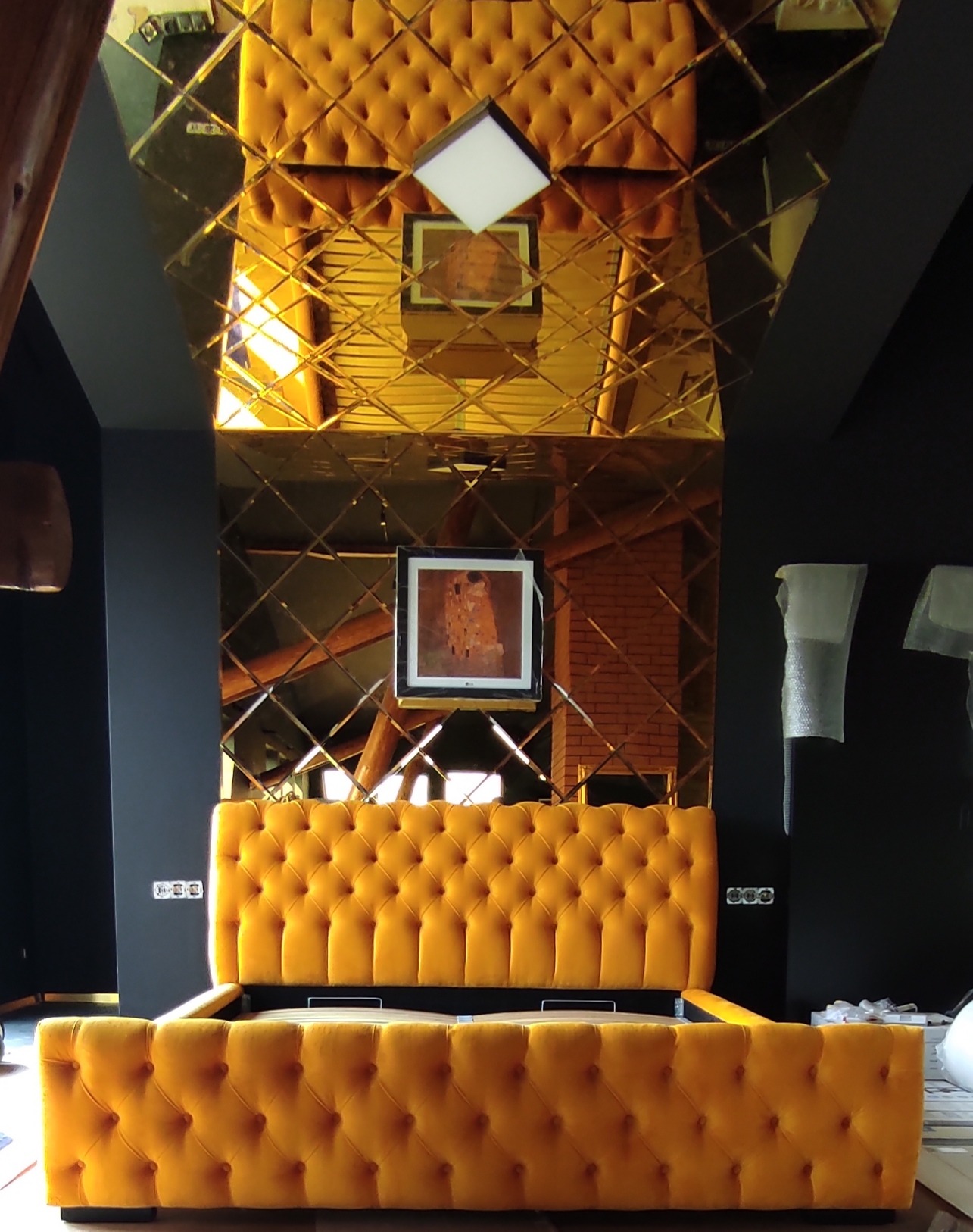 Żółte, tapicerowane łóżko z pikowanym zagłówkiem w nowoczesnym wnętrzu z lustrzanym sufitem, na którym odbija się obraz.