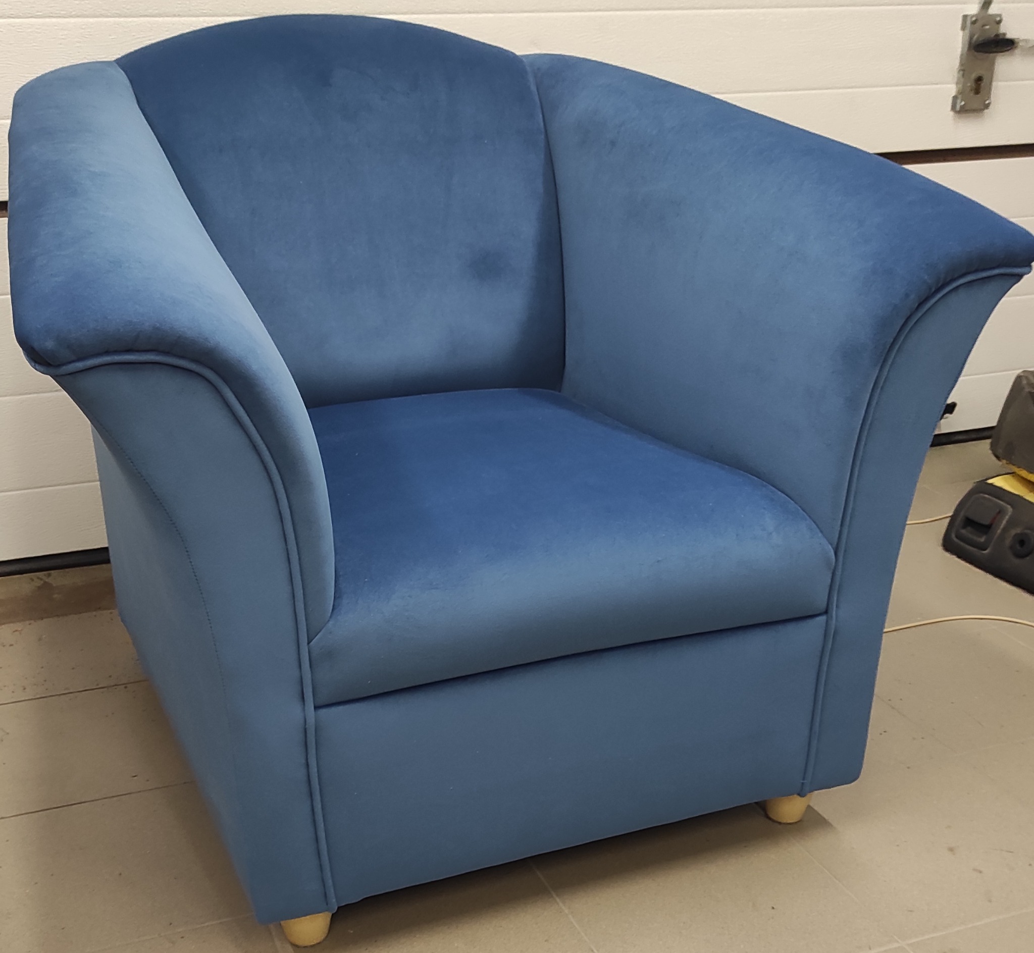 Niebieski fotel klubowy z weluru na drewnianych nóżkach, sfotografowany na tle białej bramy garażowej i narzędzi.