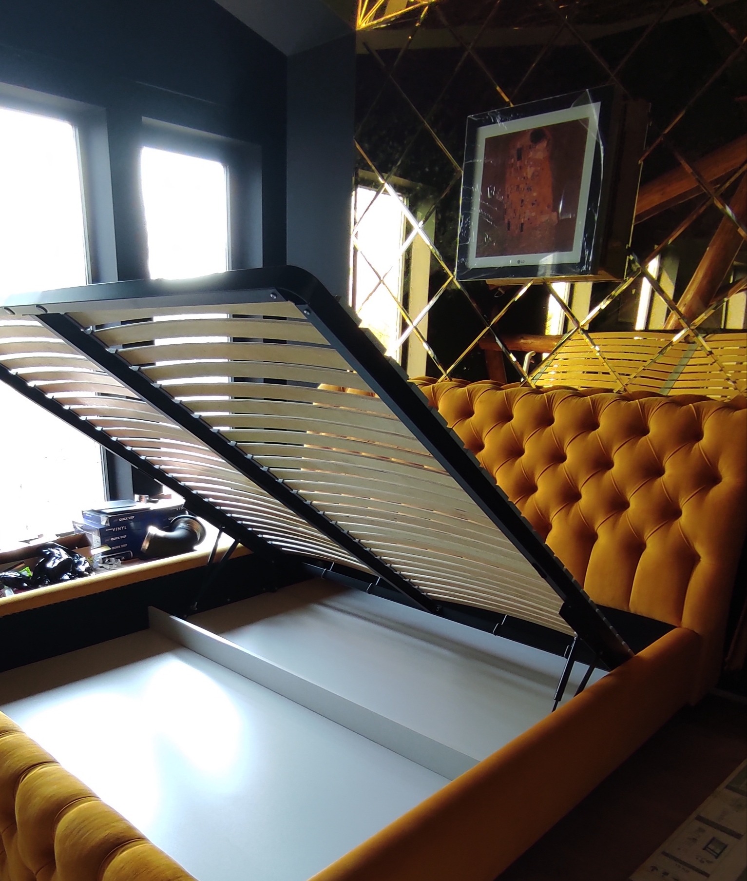 Żółte łóżko z podnoszonym stelażem i pojemnikiem na pościel w pokoju z lustrzaną ścianą i obrazem 'Pocałunek' Gustava Klimta.