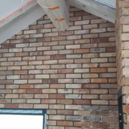 Wnętrze poddasza z fragmentem ściany wykończonej dekoracyjną cegłą w odcieniach brązu i beżu, widoczny fragment okna oraz elementy konstrukcji dachu z drewnianą belką.