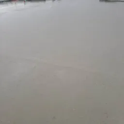 Świeżo wylana, gładka płyta fundamentowa z widocznymi elementami szalunku i zabezpieczeniem folią, w tle zabudowa jednorodzinna.